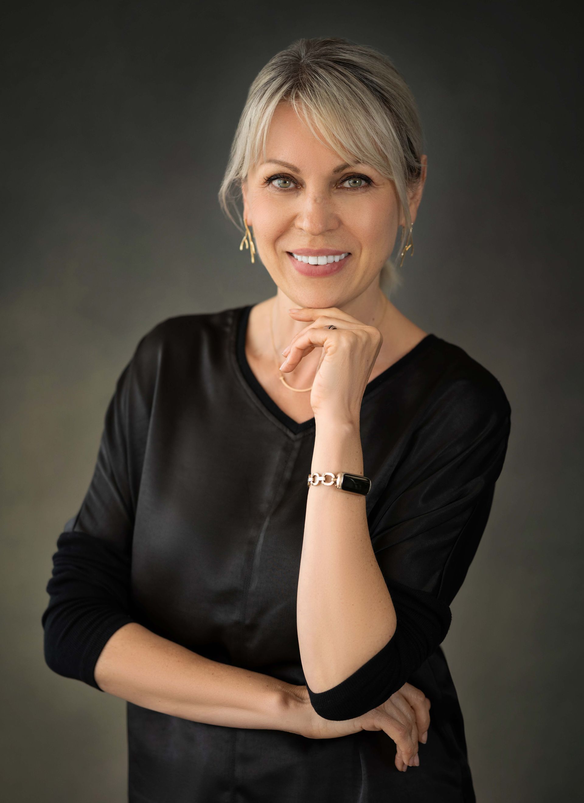 Woman with blonde hair, in black shirt, smiles with hand under chin against gray background.