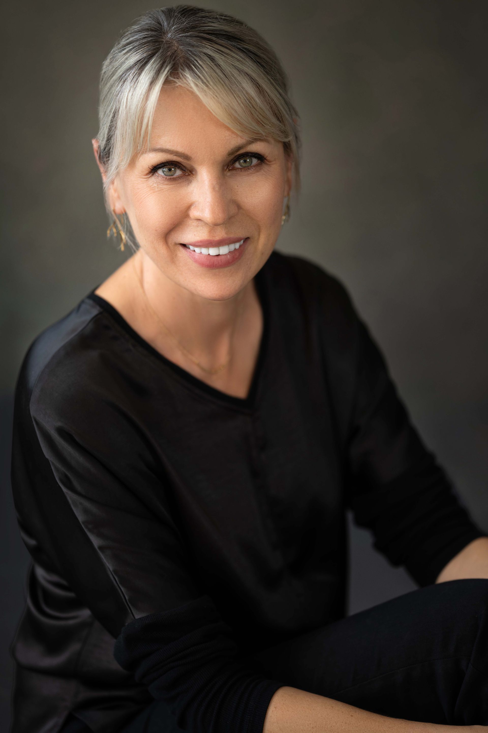 Woman with short blonde hair smiles, wearing a black top against a gray backdrop.