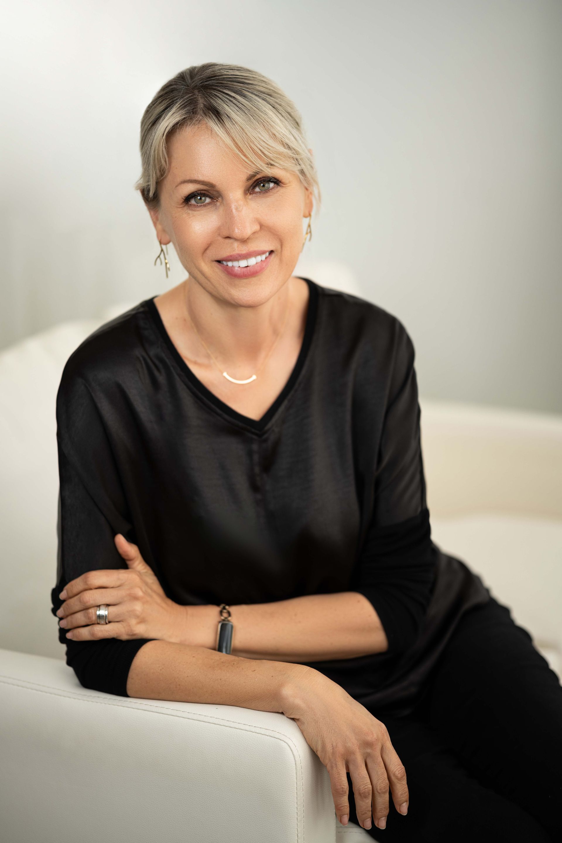Woman with blonde hair smiles while seated on a white couch, wearing a black top and jewelry.