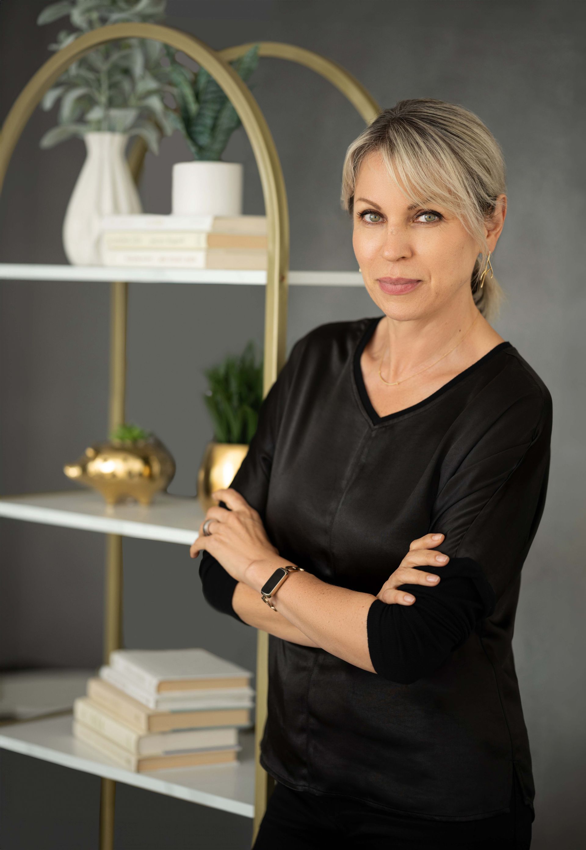 Woman with crossed arms, in a black top, stands in front of a gold bookshelf.
