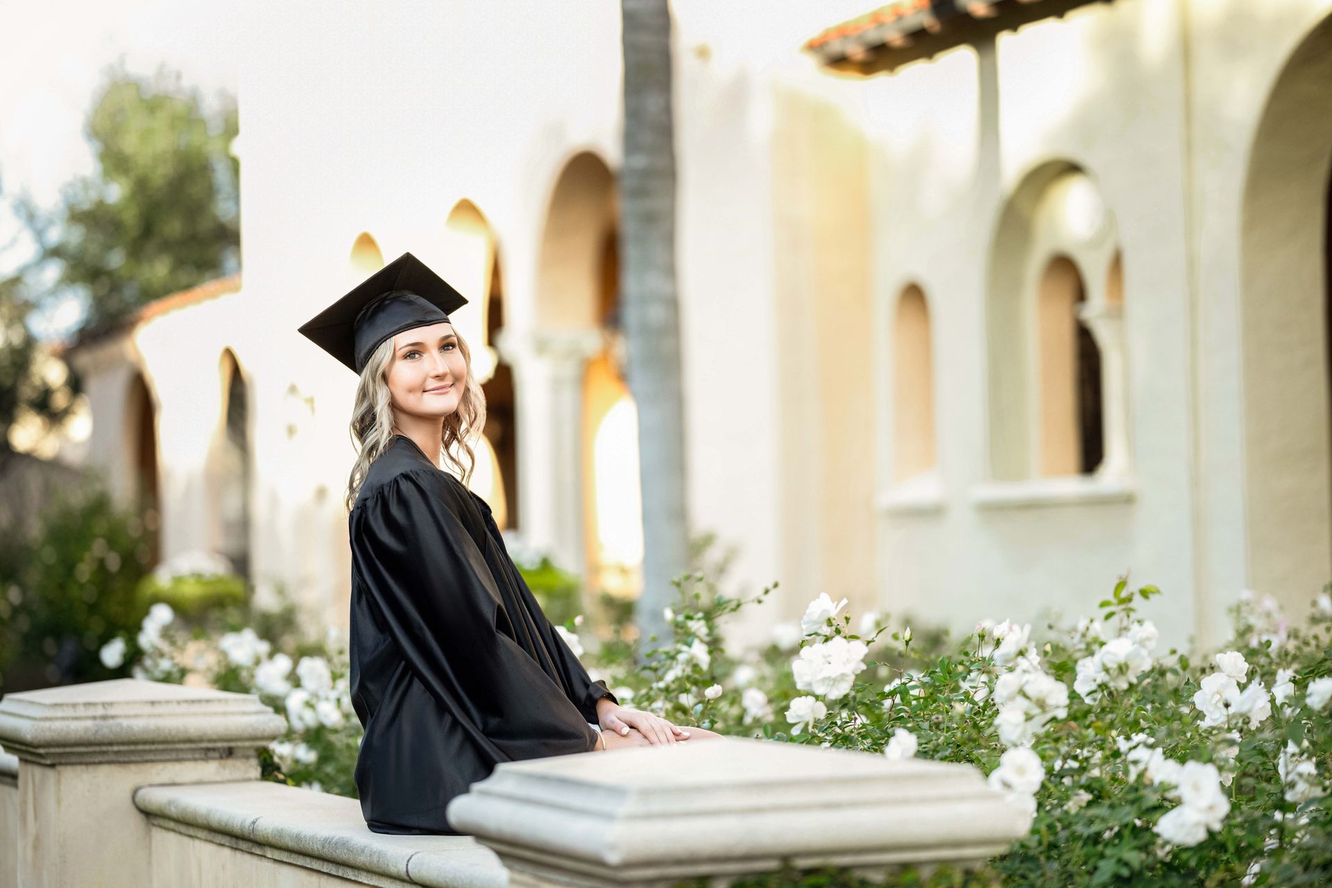 Woman in graduation cap and gown smiles in front of building with white roses.