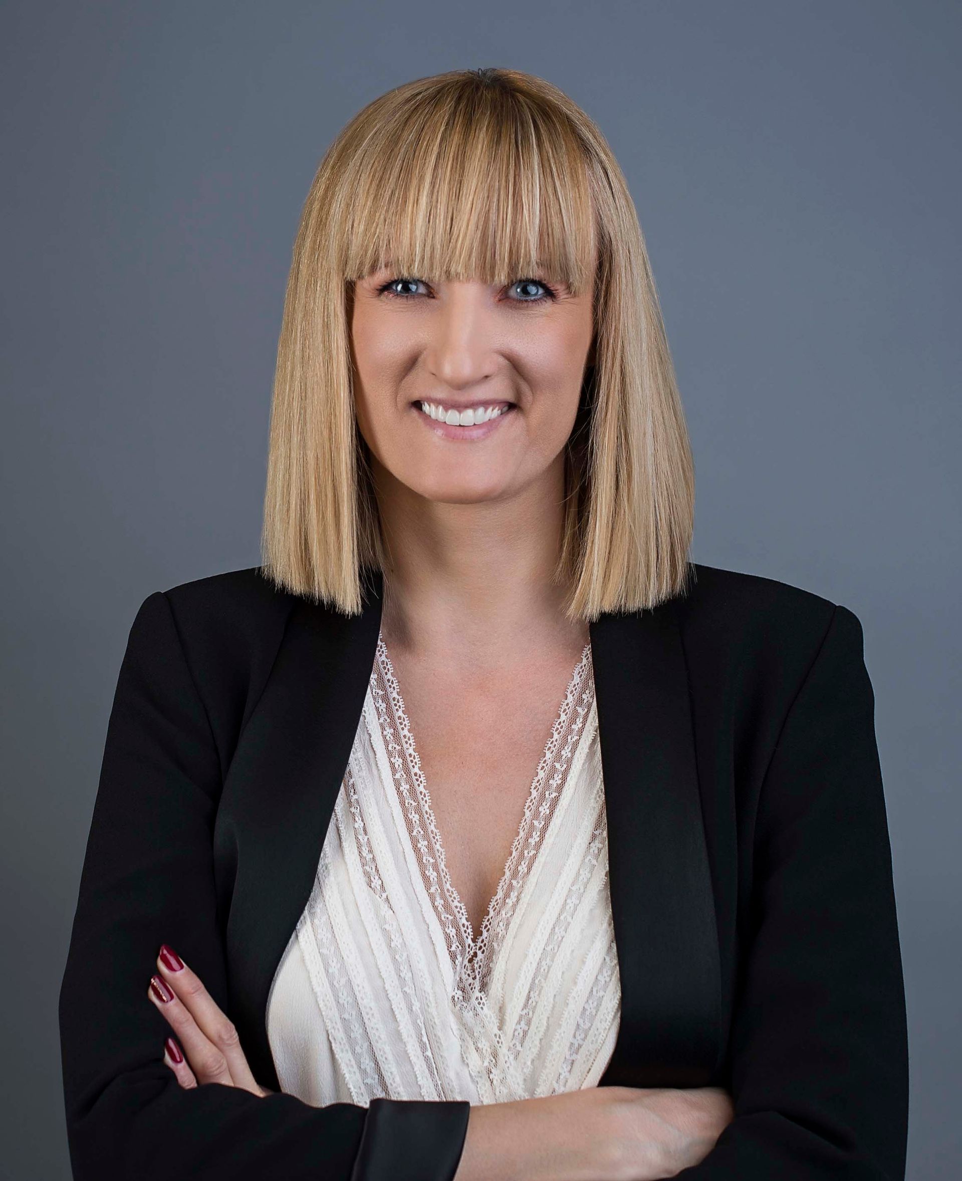 Woman with blonde hair, black blazer, white shirt, arms crossed, smiles. Gray background.