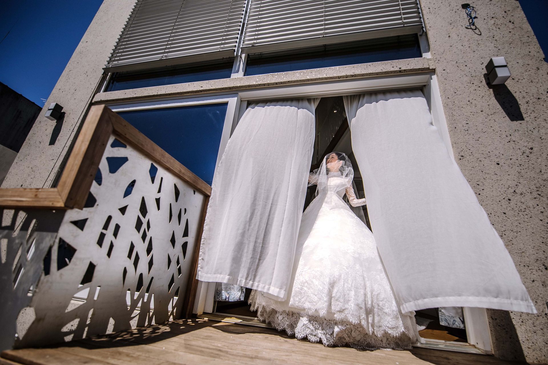 Bride in a white wedding dress stands in doorway, curtains blowing, sunlight, modern building exterior.