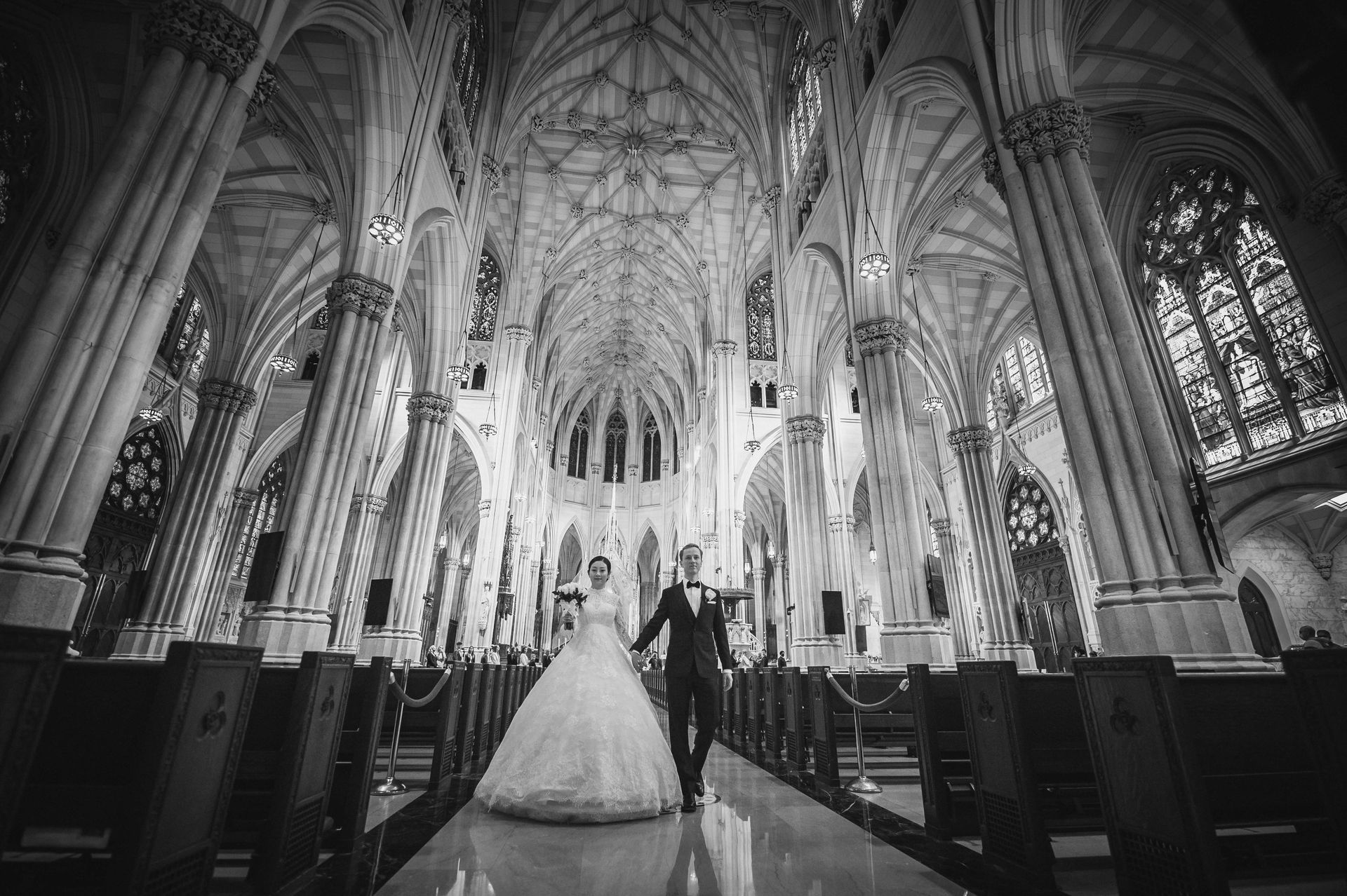 Bride and groom hold hands, walking down aisle in ornate cathedral. Black and white.