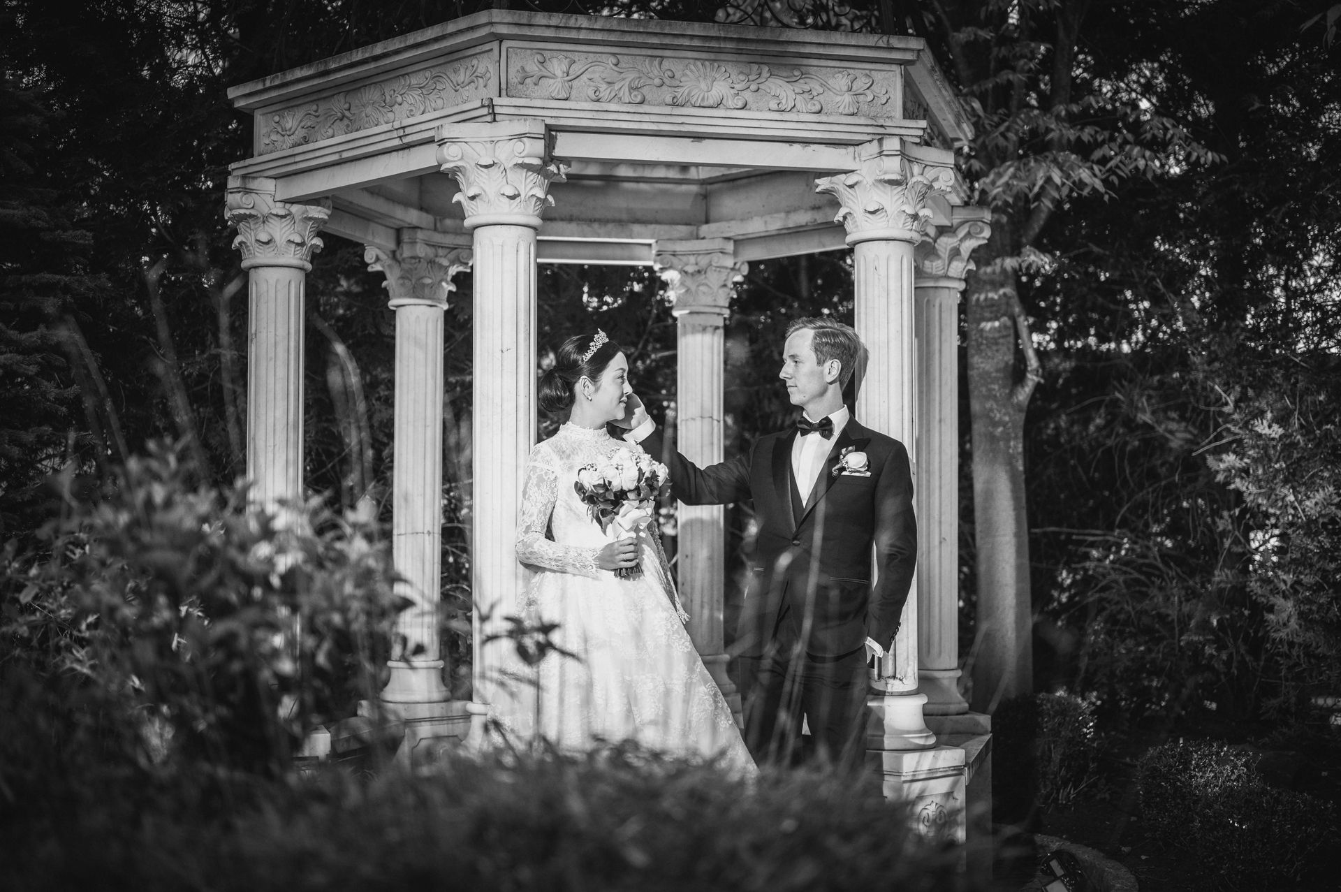 Bride and groom stand under a gazebo. The groom touches the bride's shoulder. Black and white photo.