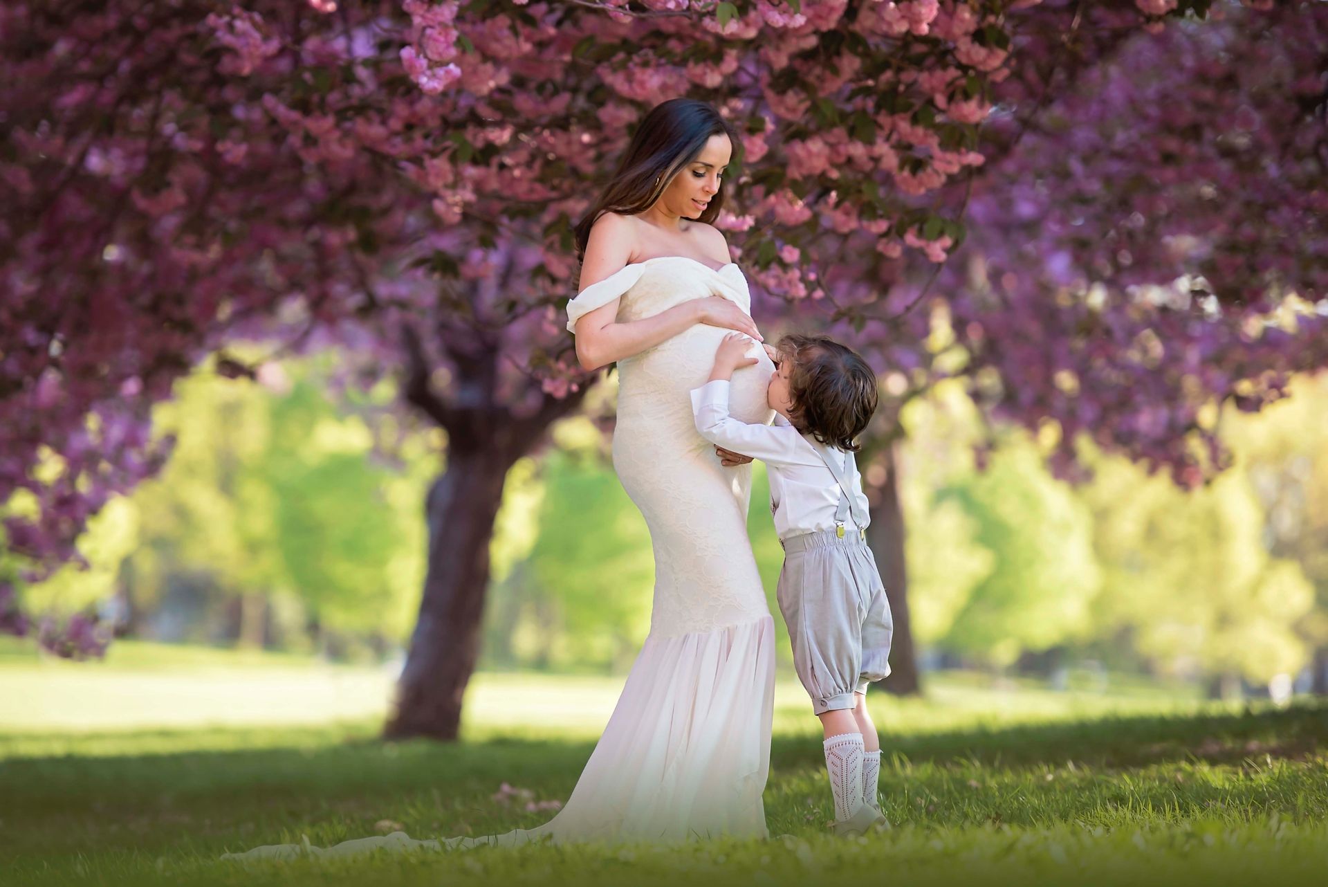 Pregnant woman in white gown and child under flowering tree in a park.