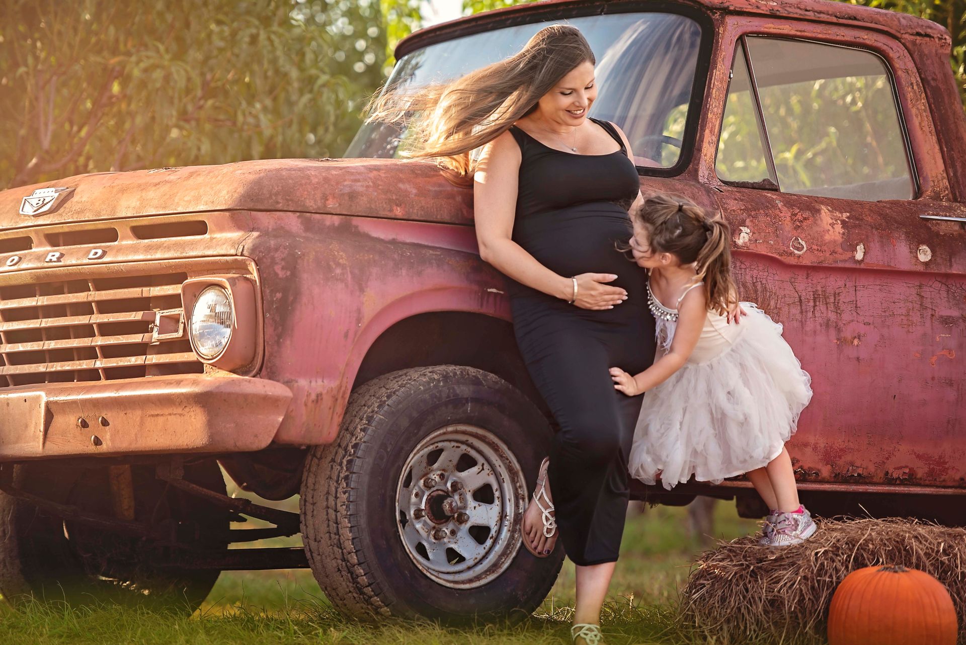 Pregnant woman and young girl by an old red truck. The girl kisses the woman's belly. Golden hour.