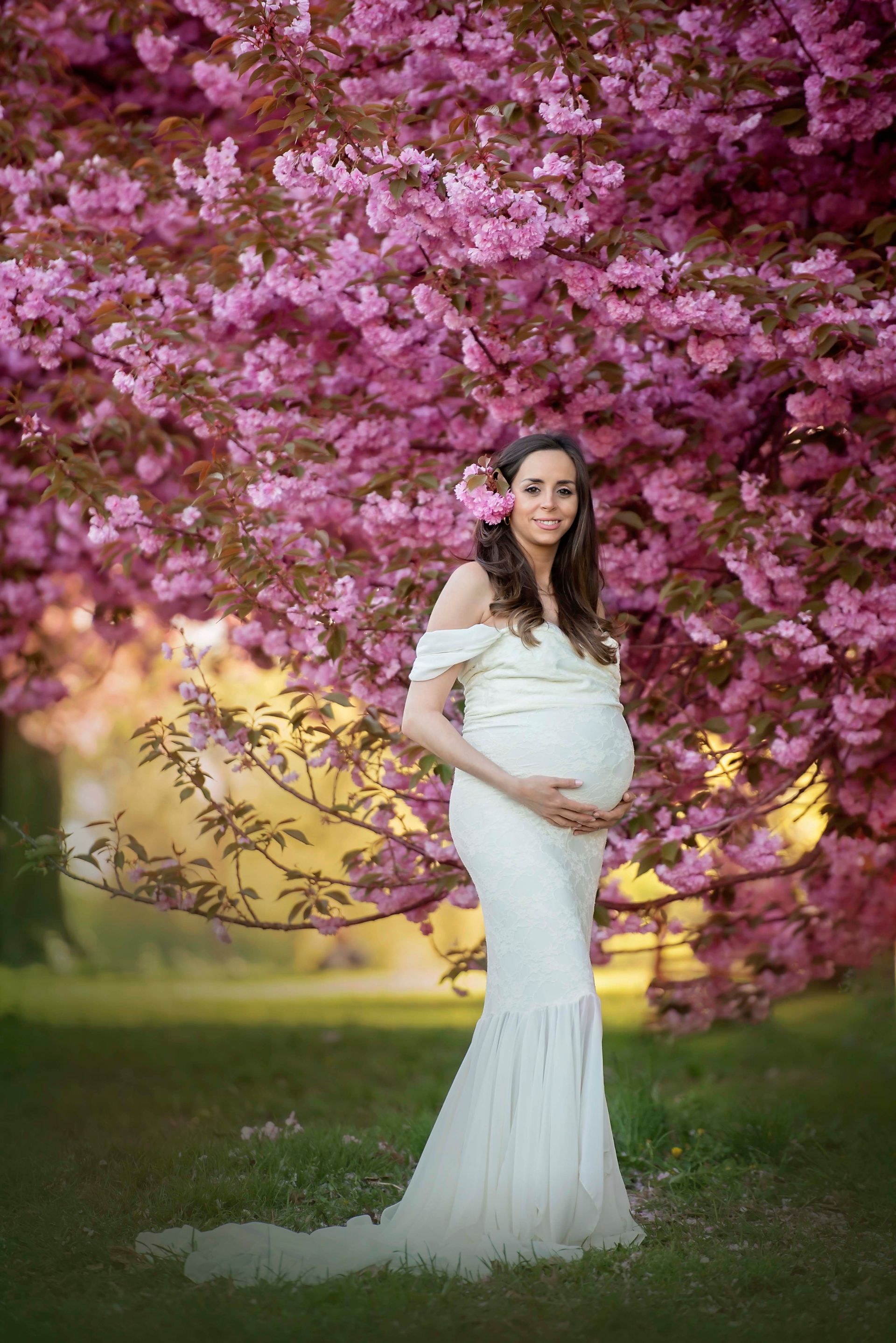 Pregnant woman in white gown poses in front of a blooming pink tree, holding her belly.