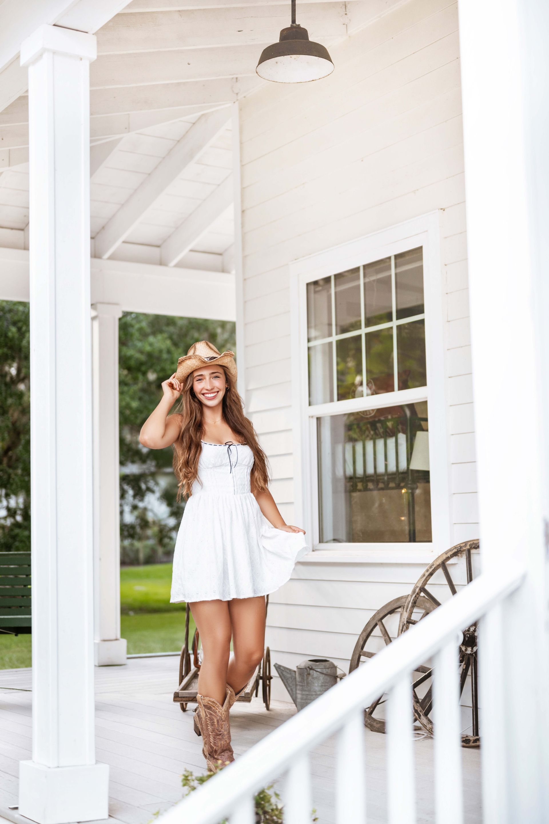Woman in cowboy hat and boots smiles on a white porch, wearing a white dress.