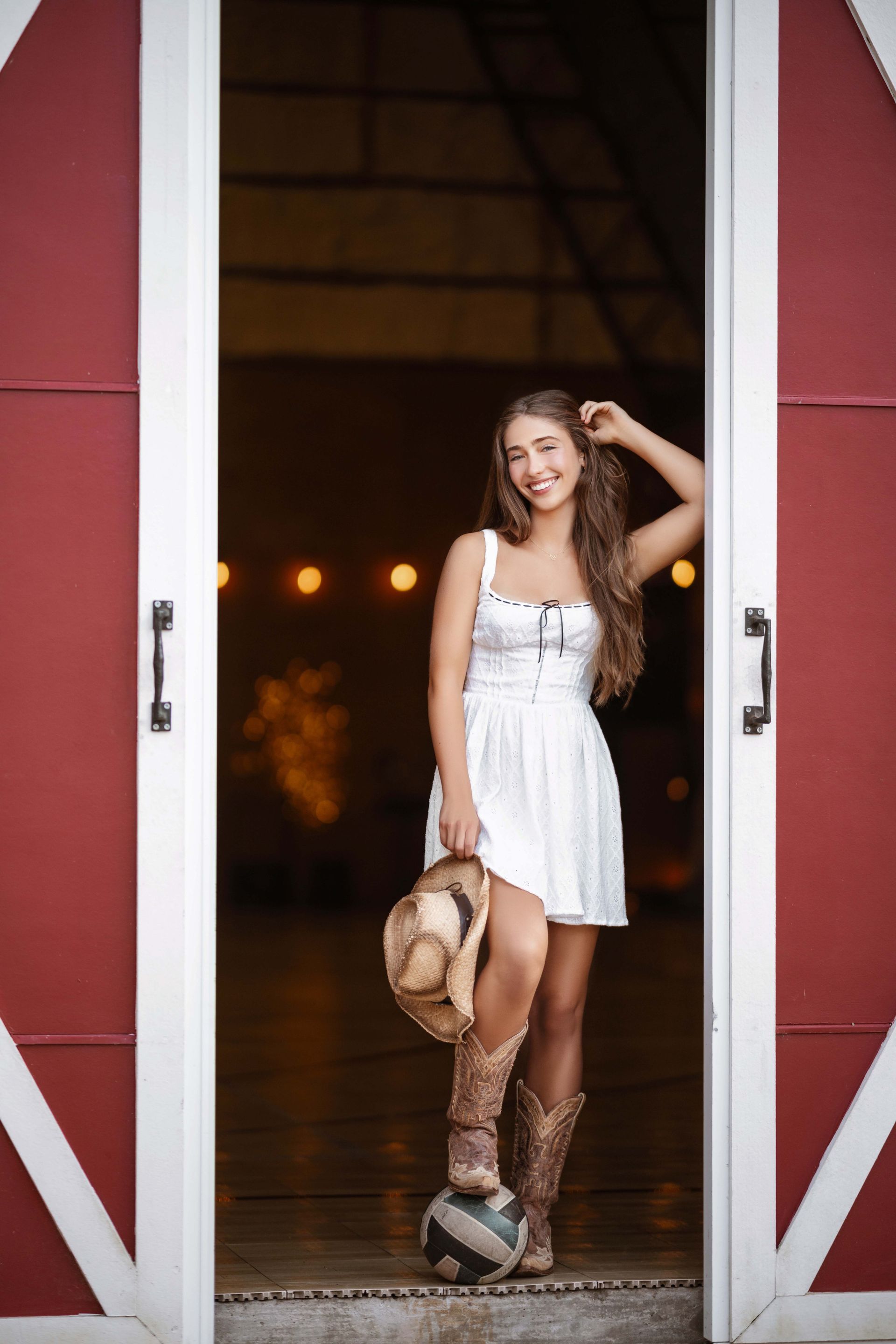 Young woman in white dress and boots leans in barn doorway, smiling and holding a hat.