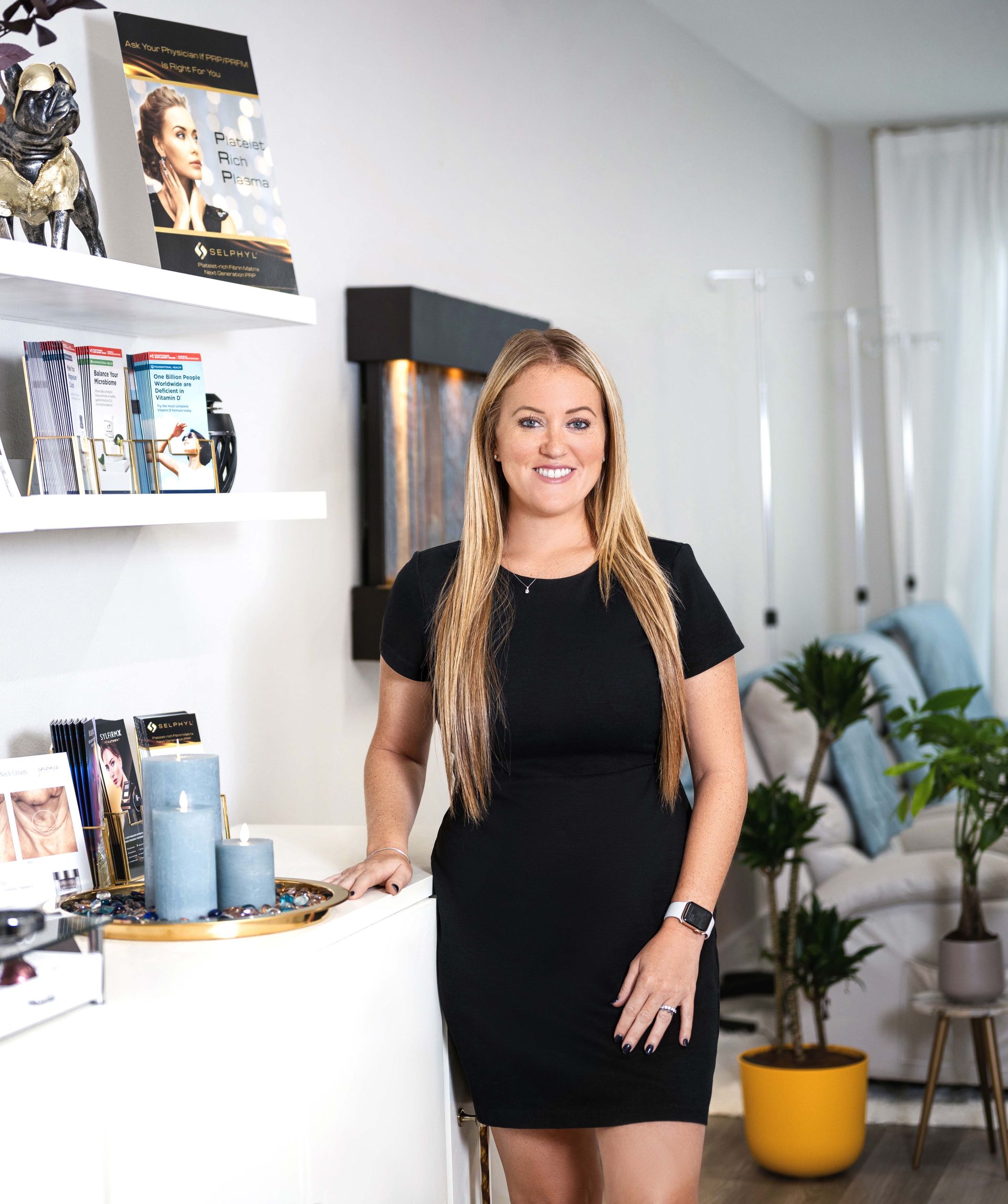 Woman in black dress, standing in room, smiling. White shelves with books and candles beside her.