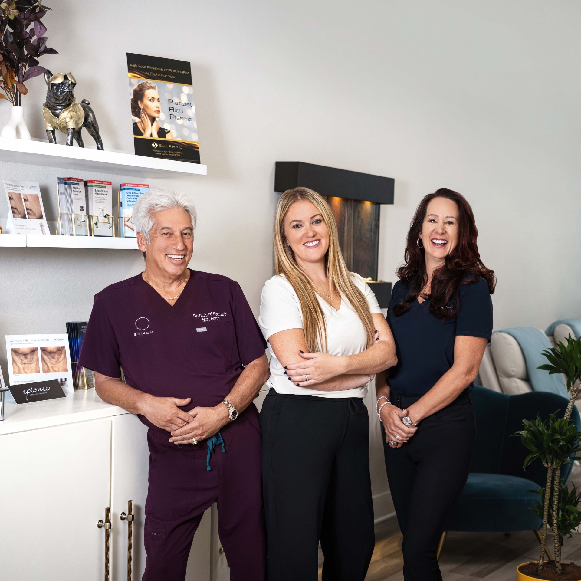 Three smiling people in a clinic: A doctor in purple scrubs, a woman in a white top, and a woman in a dark blue top.