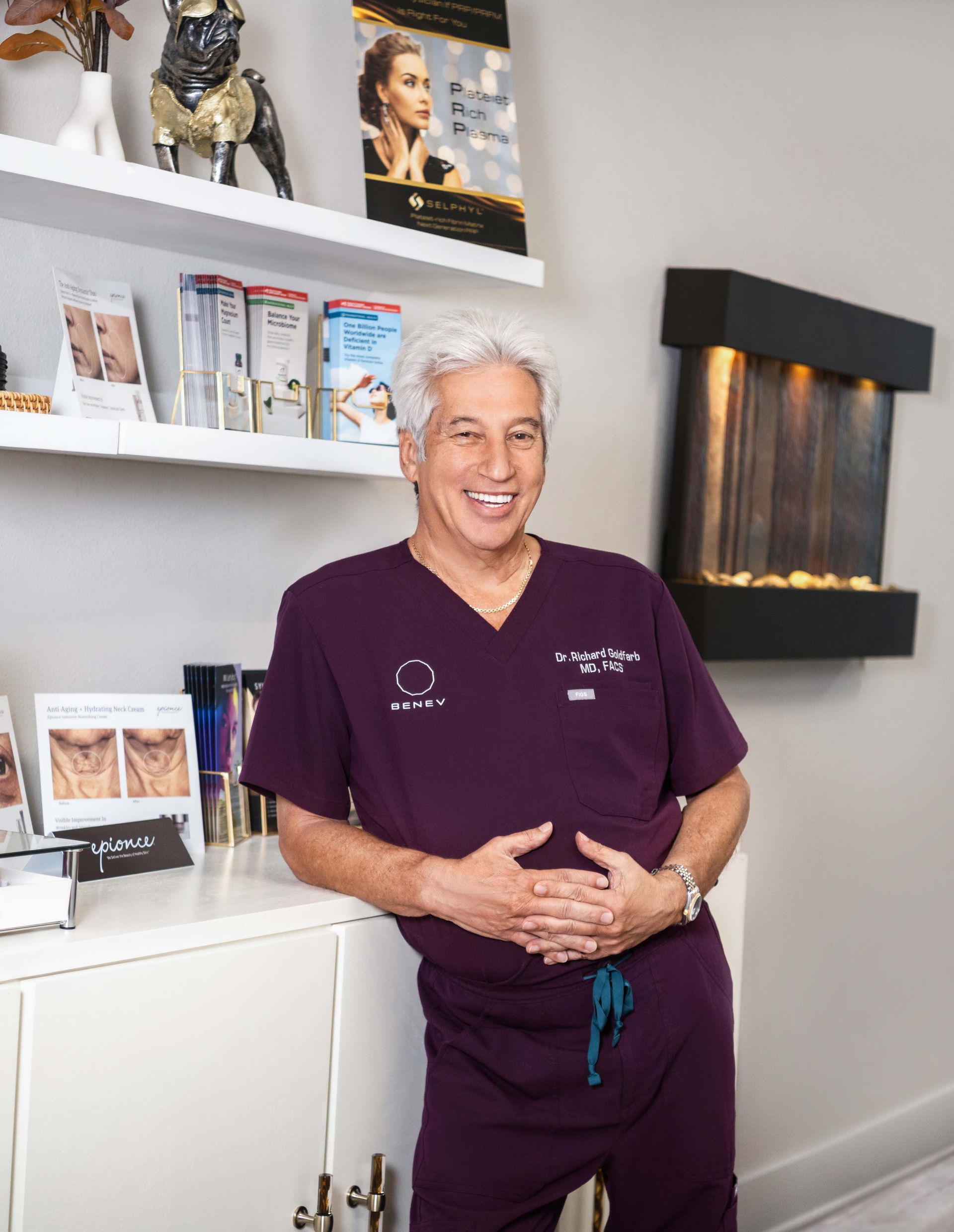 Man in purple scrubs smiles, leaning against a cabinet in a medical office.