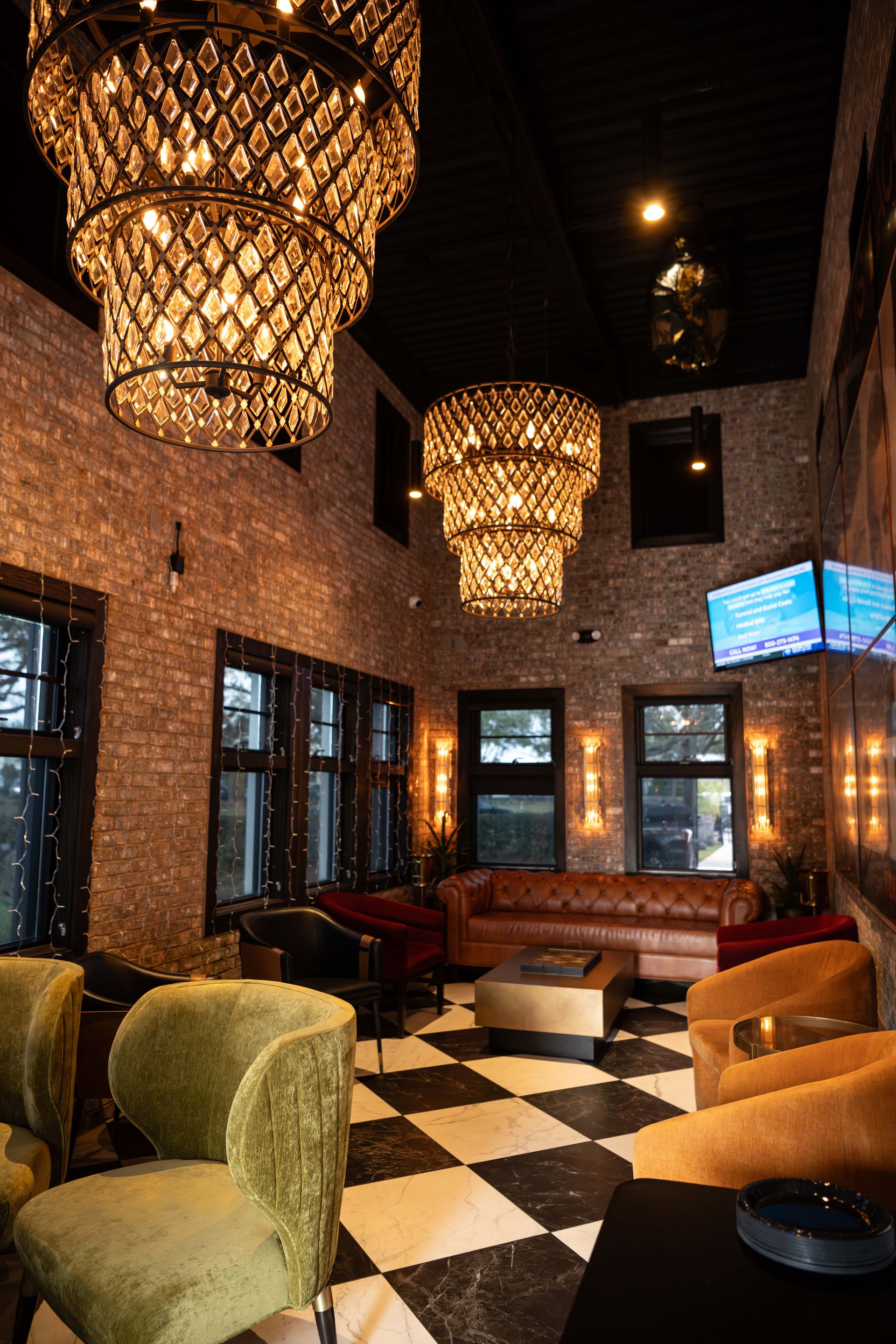 Elegant lounge with brick walls, chandeliers, and checkered floor. Furnished with couches and velvet chairs.