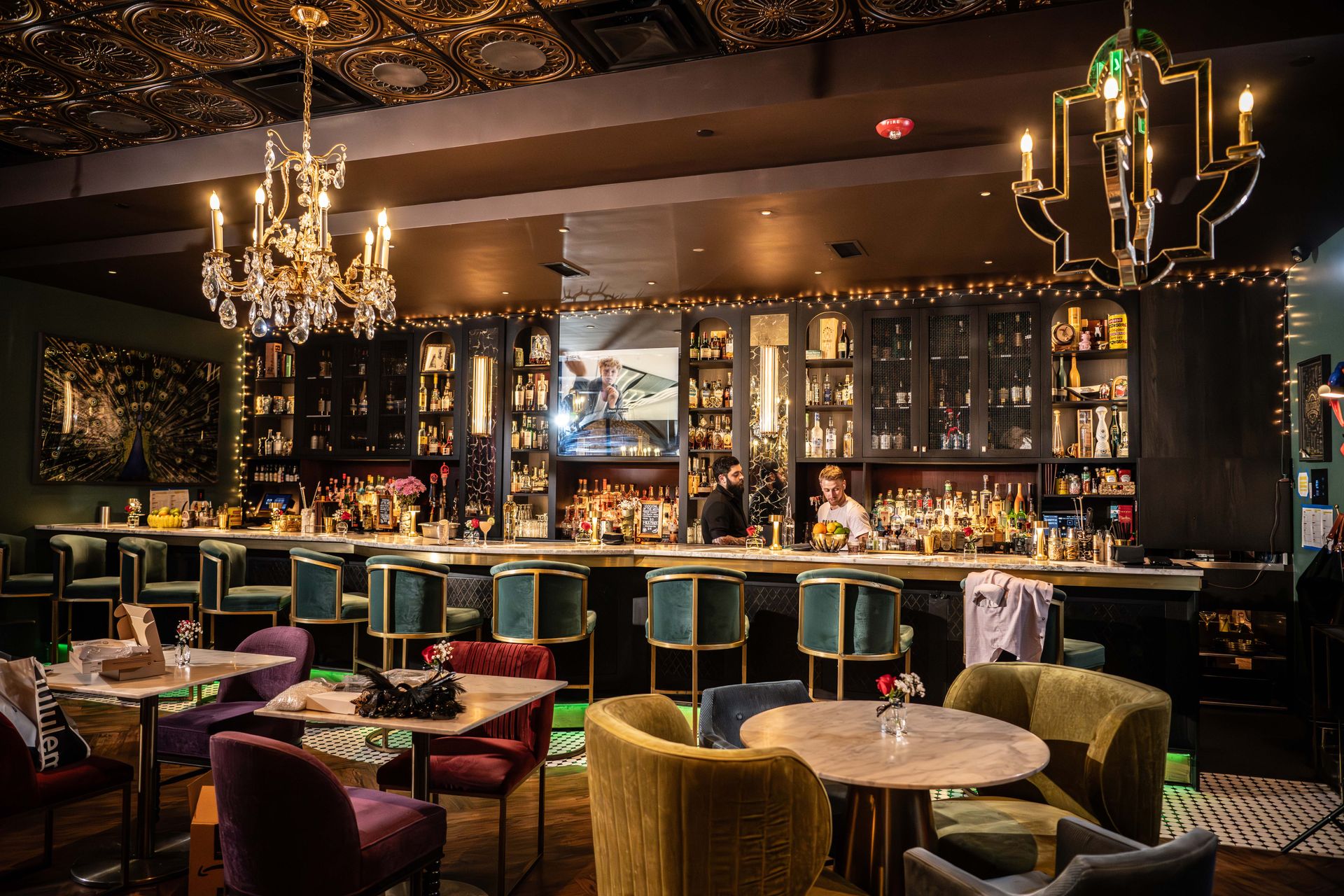 An elegant bar interior with jewel-toned seating, chandeliers, and bartenders at work.