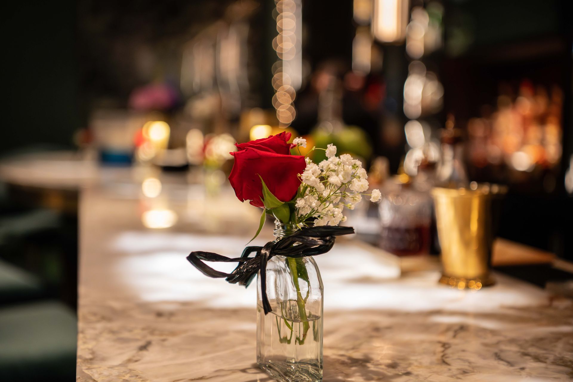 Red rose and white flowers in a small glass vase on a marble bar, with blurred bar background.