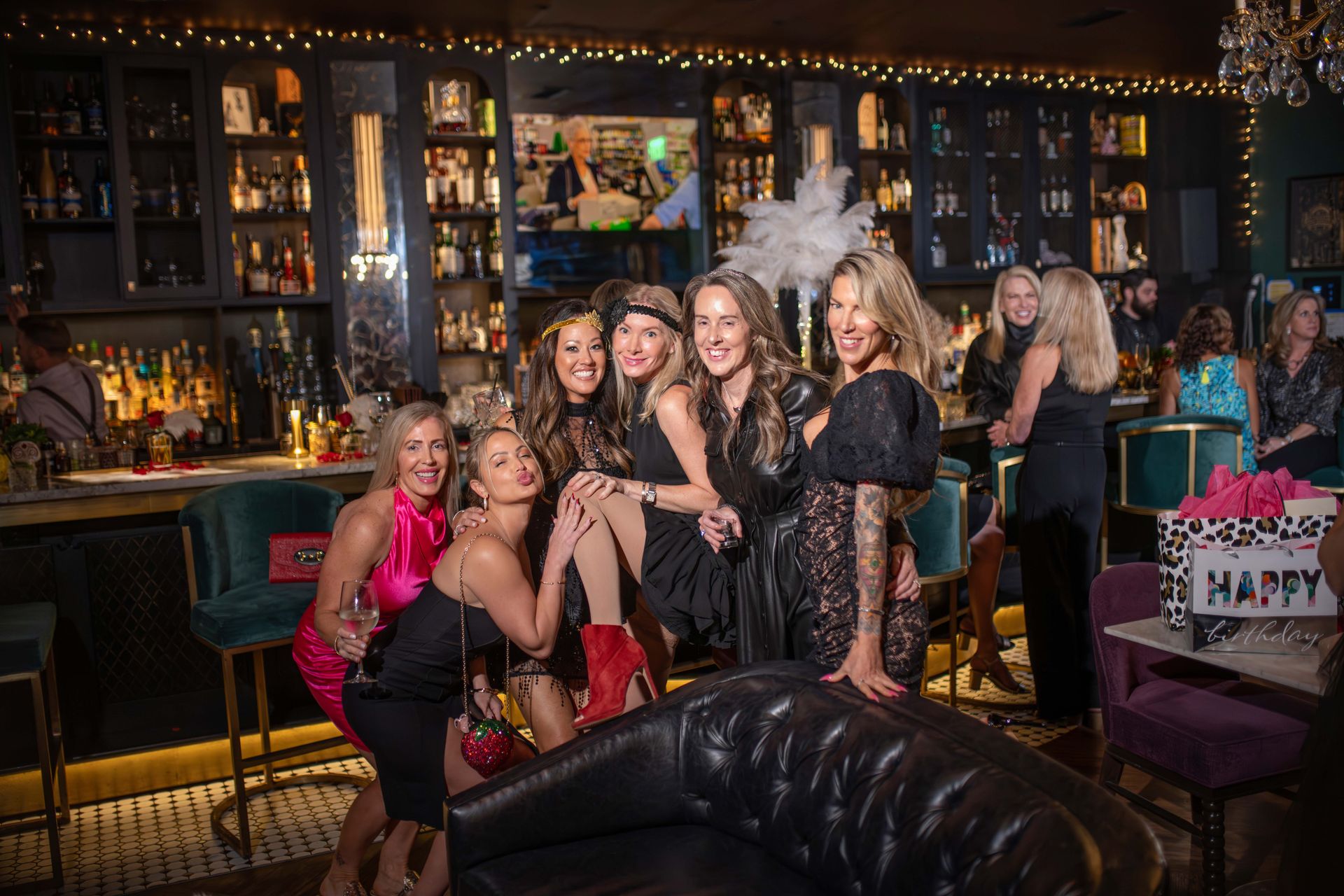 Group of women posing at a bar. One is being held up. Dark decor, festive atmosphere.
