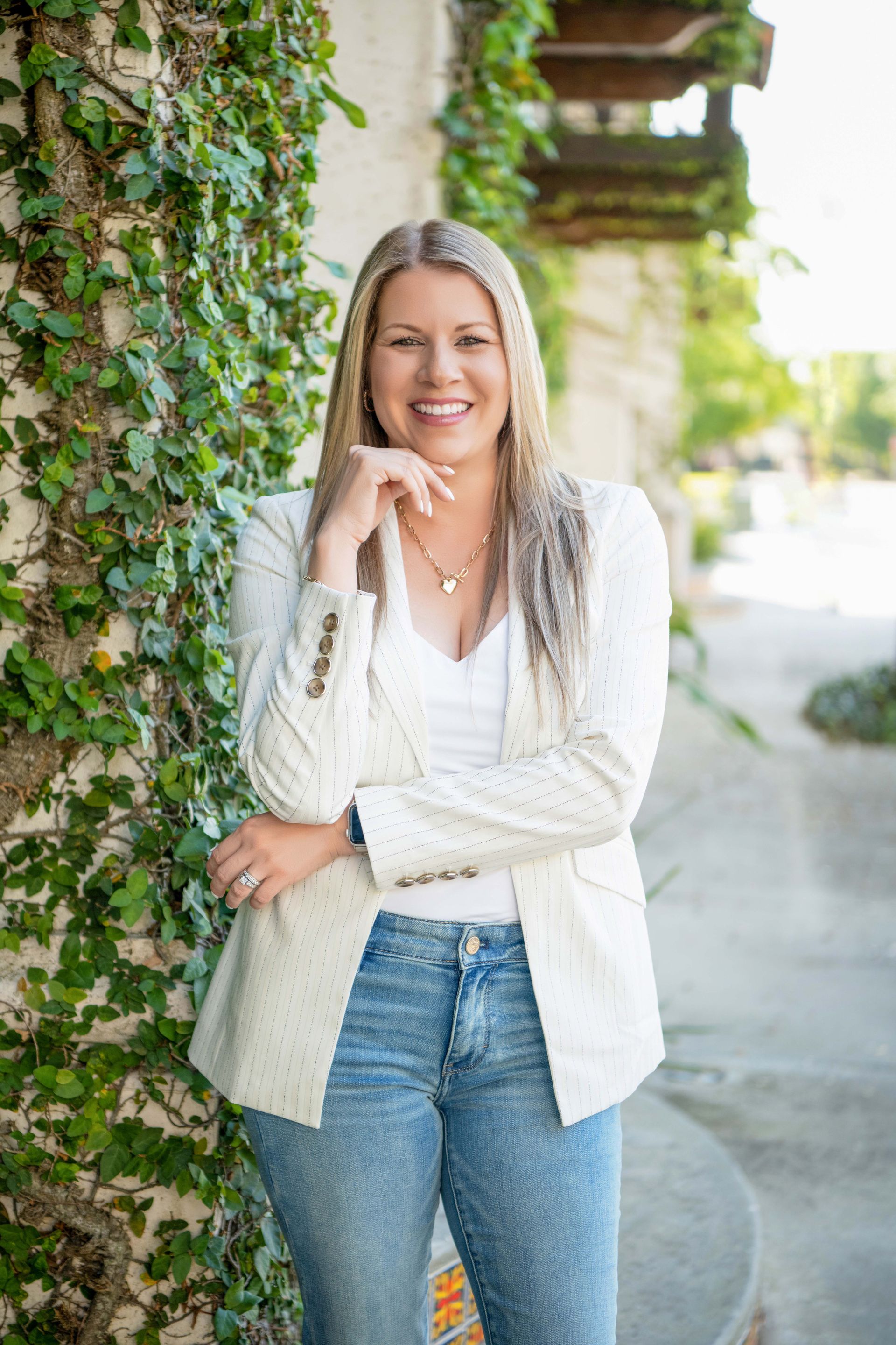 Woman with blonde hair, in a white blazer and jeans, smiles, resting chin on hand, standing near a vine-covered wall.