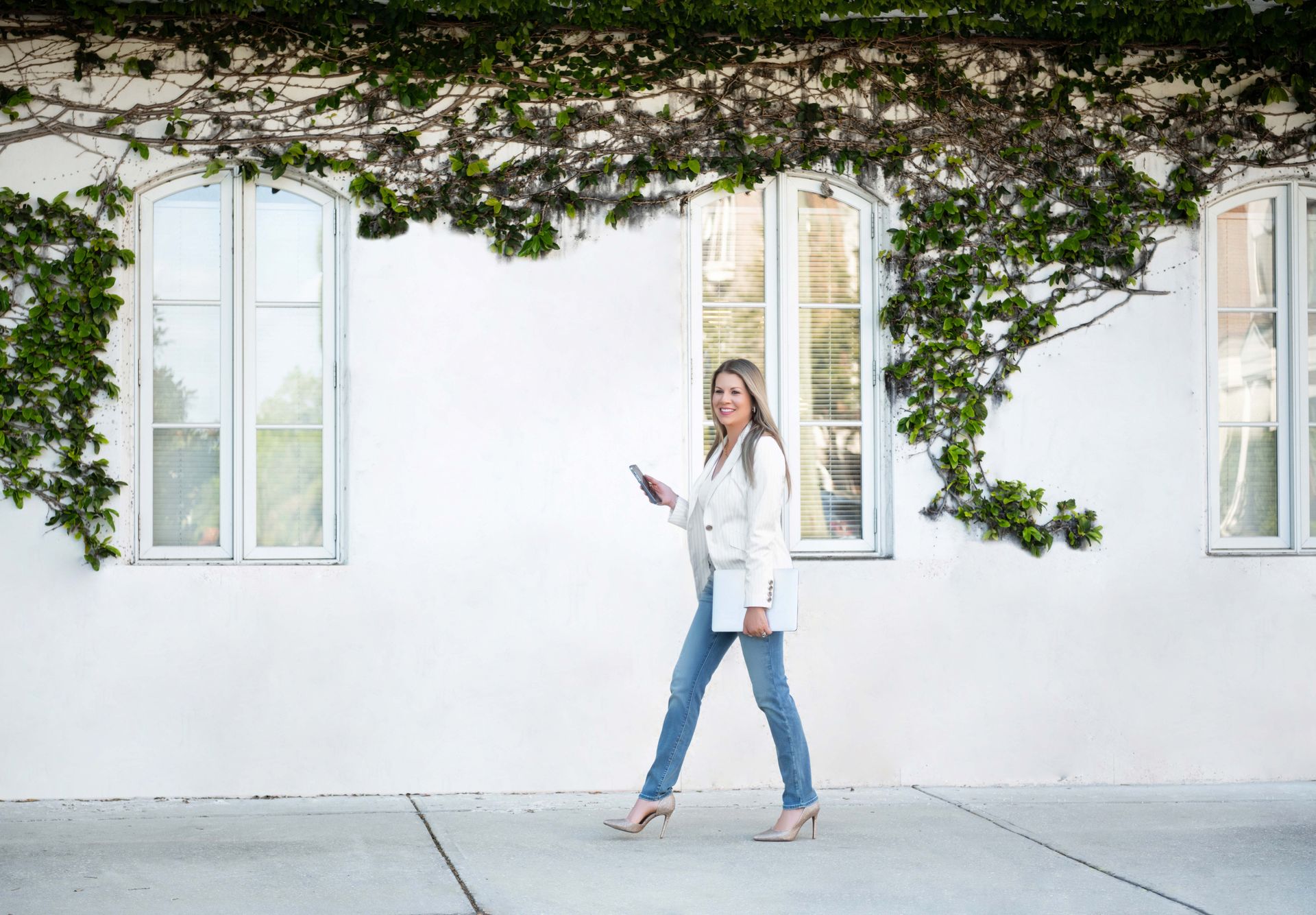 Woman in white blazer, jeans, and heels walks on a sidewalk, looking at her phone, with ivy on the wall behind her.