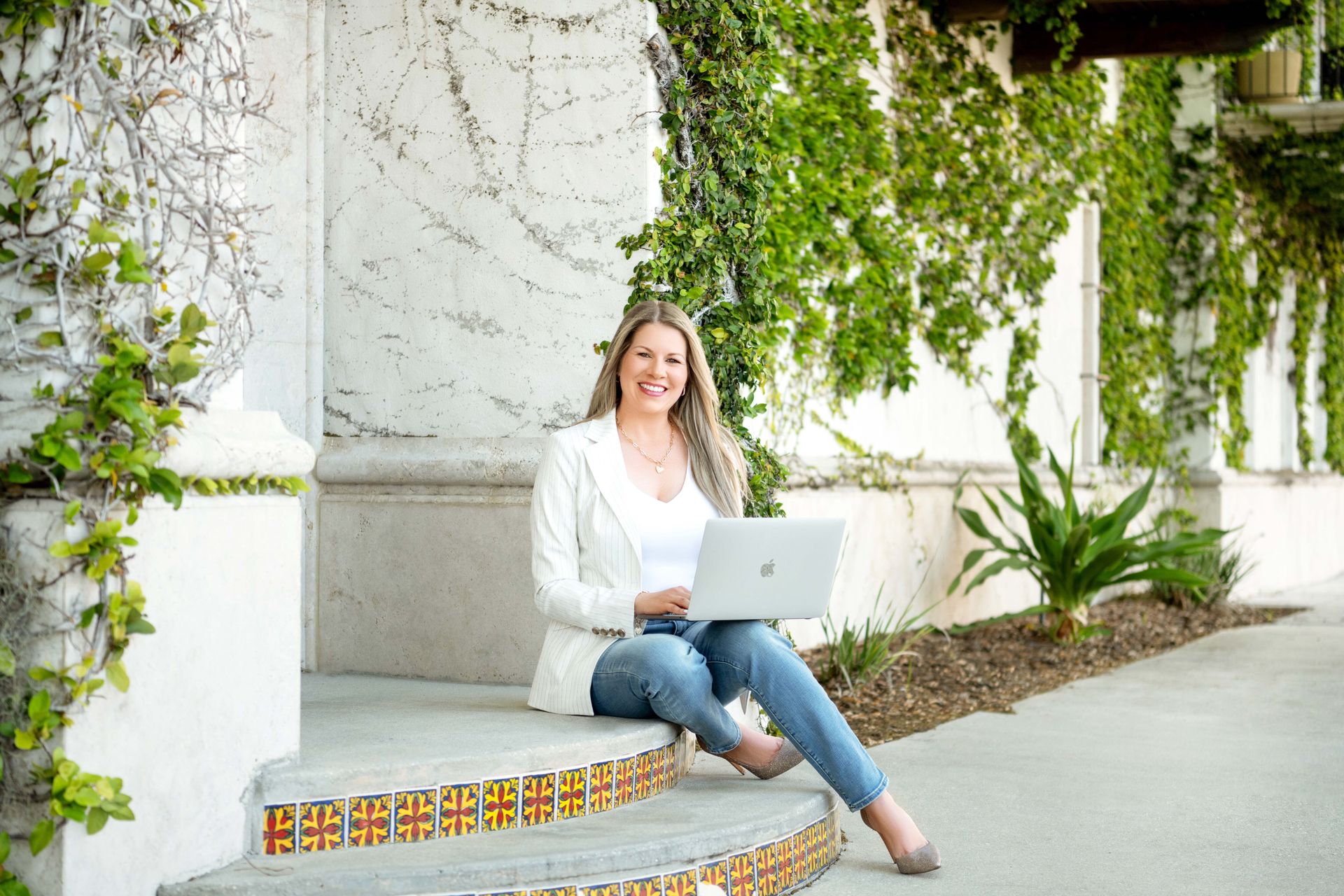 Woman sitting on steps, working on a laptop, wearing white jacket, jeans, smiling, outdoors with greenery.