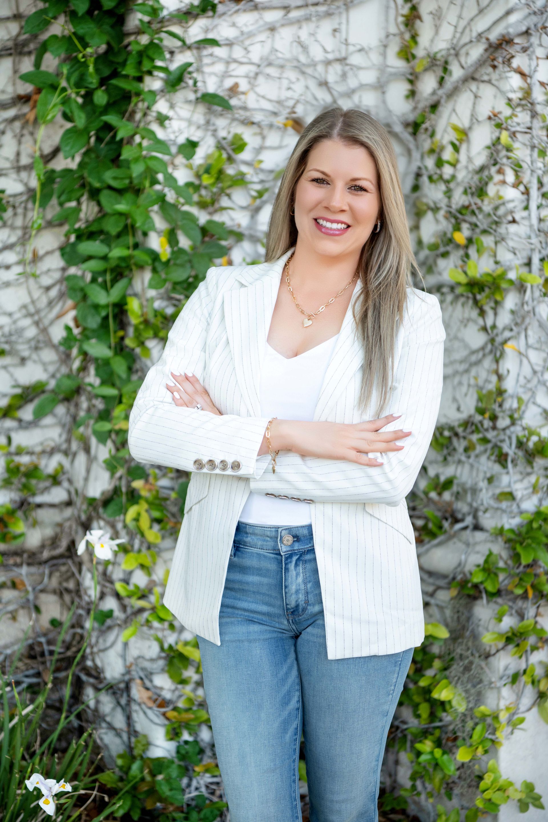 Woman smiling, wearing a white blazer and jeans, arms crossed, standing in front of a green ivy-covered wall.