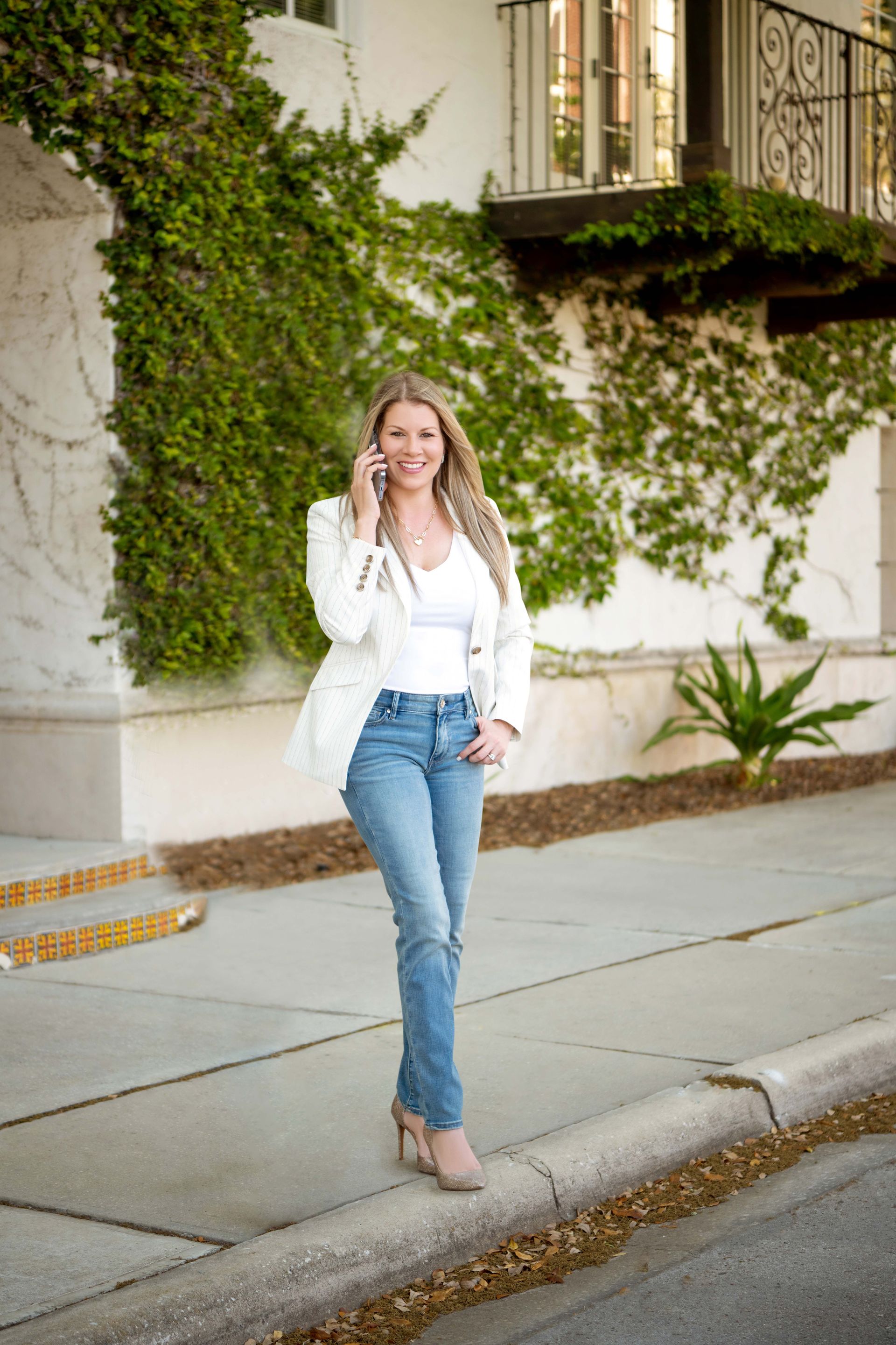 Blonde woman in white blazer, jeans, talking on phone, smiling, near a building covered in ivy.