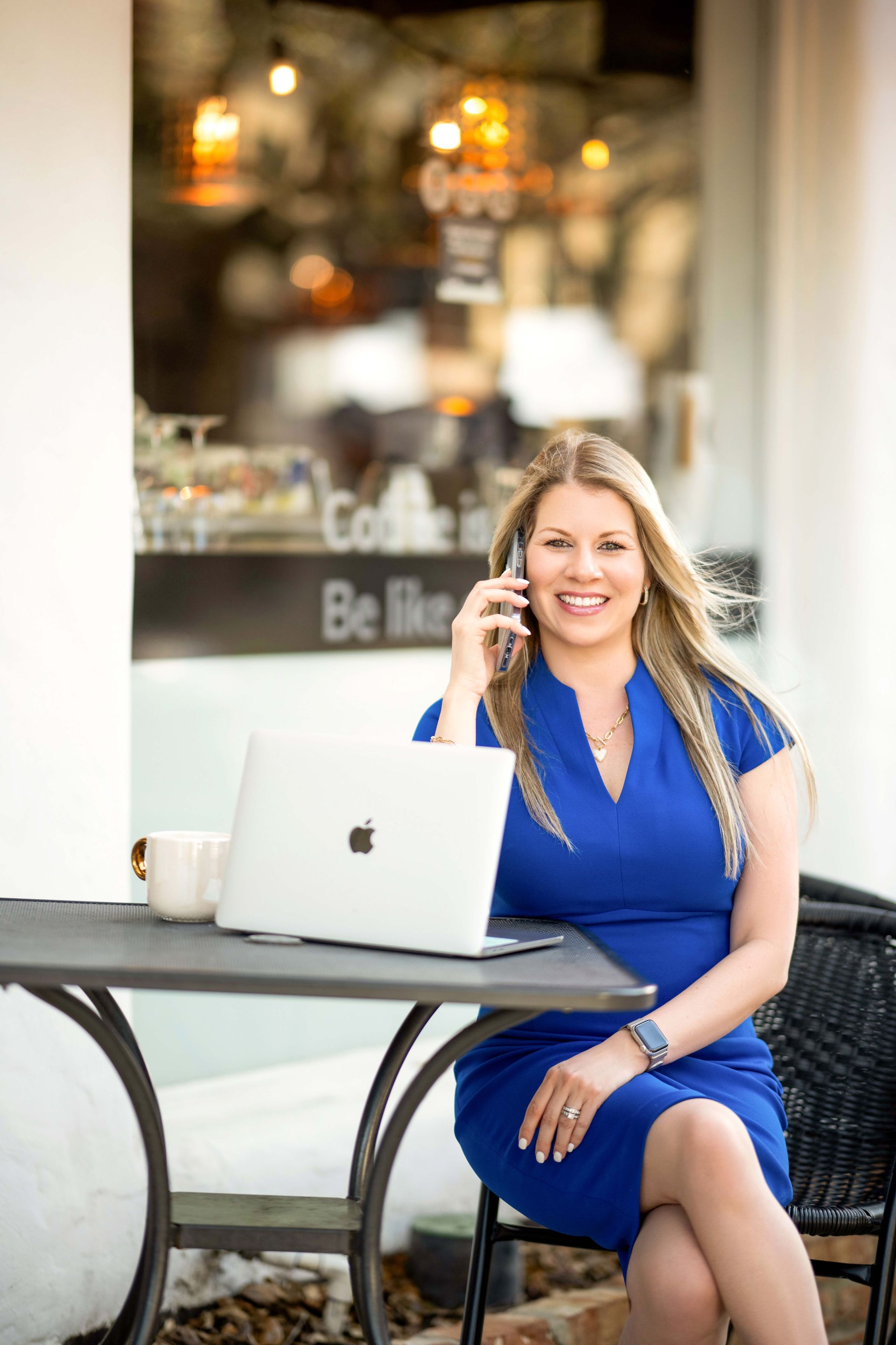 Woman in blue dress, talking on the phone, seated at a cafe table with a laptop.