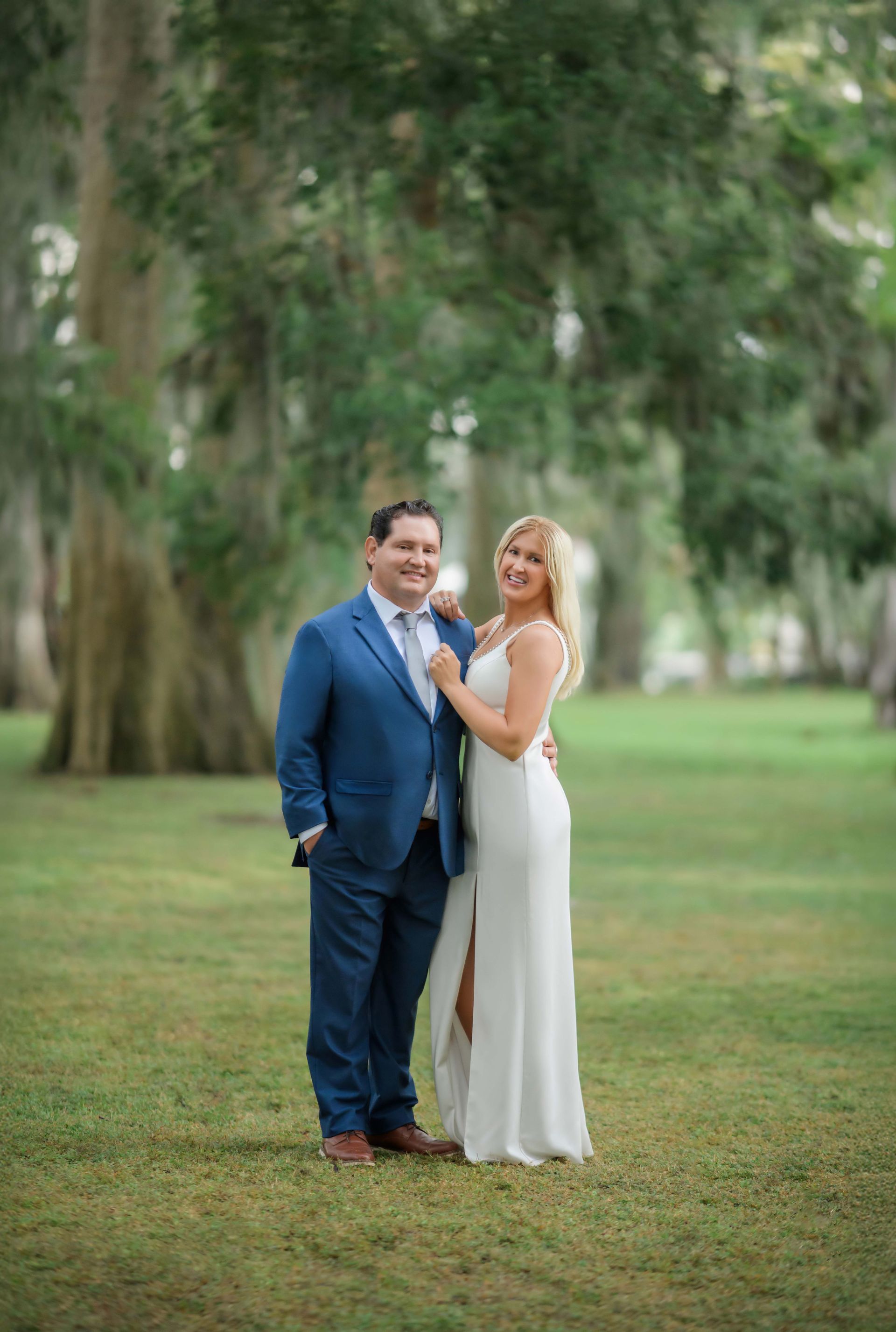 Wedding couple, man in blue suit and woman in white dress, posing outdoors in park.