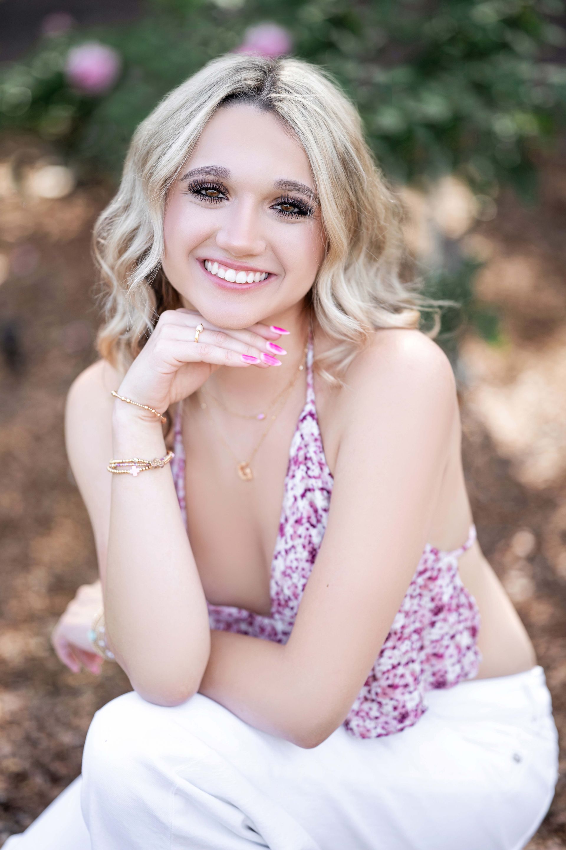 Blonde woman smiling, posing outdoors in white pants and floral top.