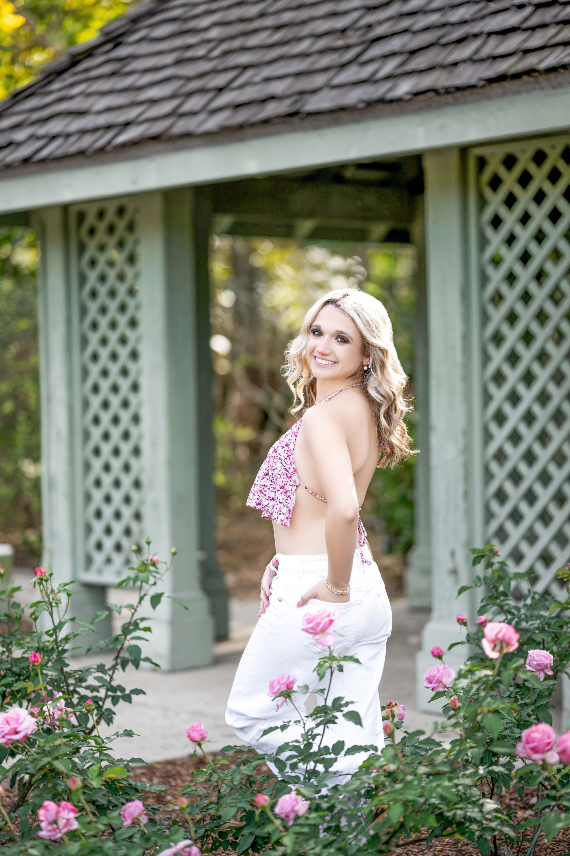 Blonde woman in pink top and white pants poses in a garden with a gazebo.