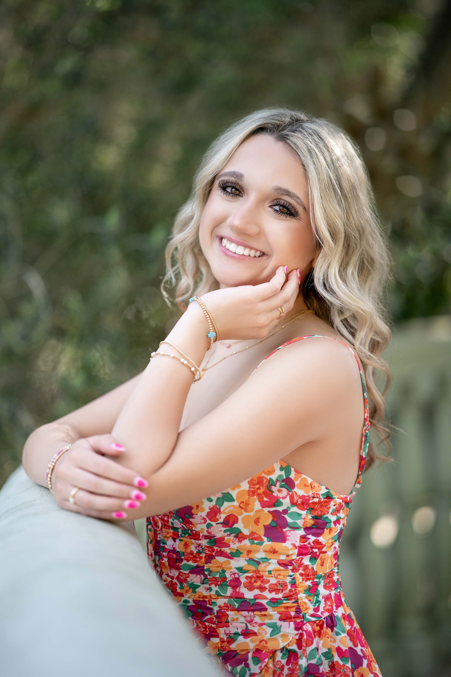 Blonde woman smiling, leaning on a railing. Wearing floral dress, bracelets. Outdoors with trees in the background.