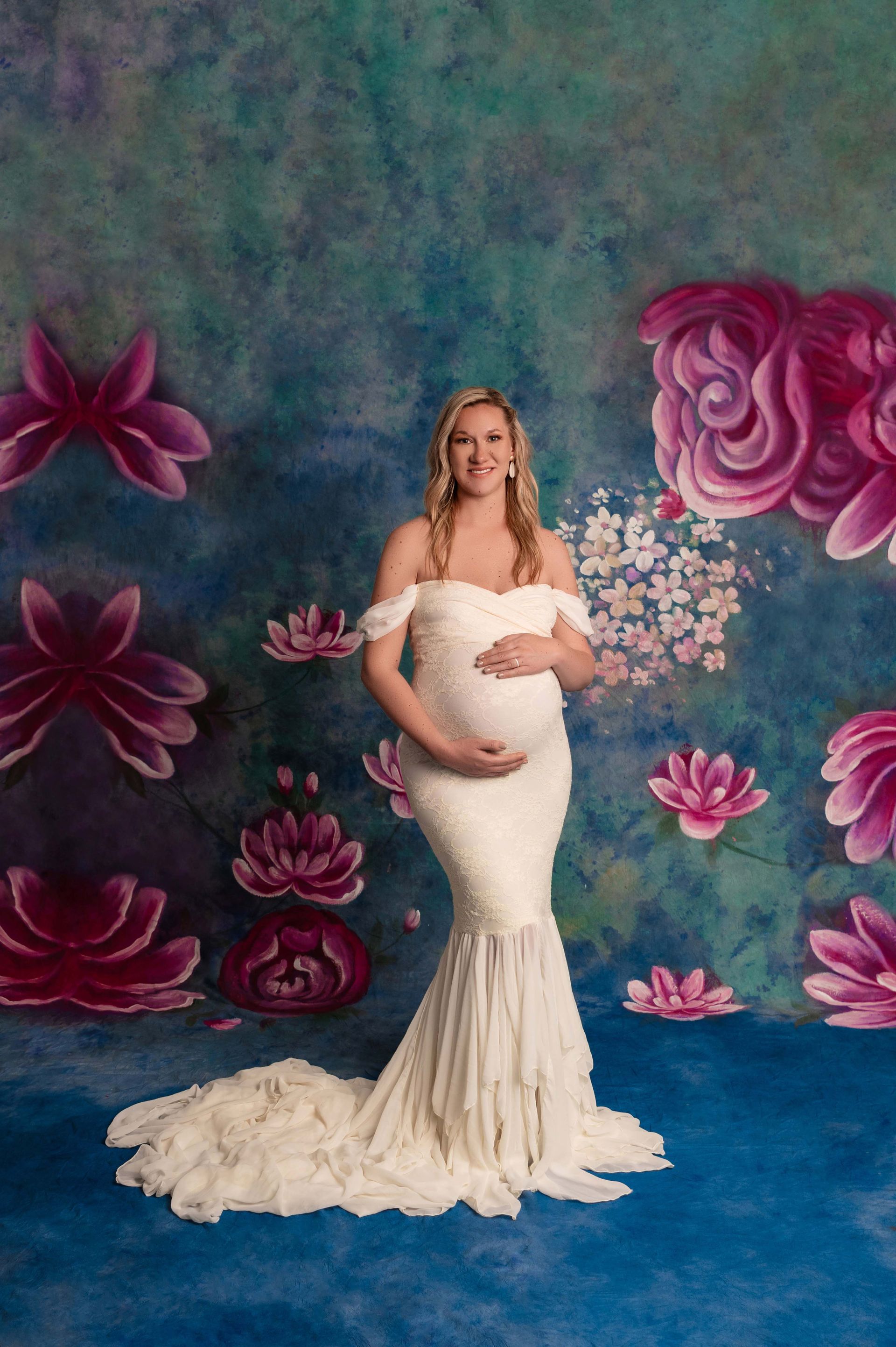 Pregnant woman in white gown, smiling, poses against a blue floral backdrop.