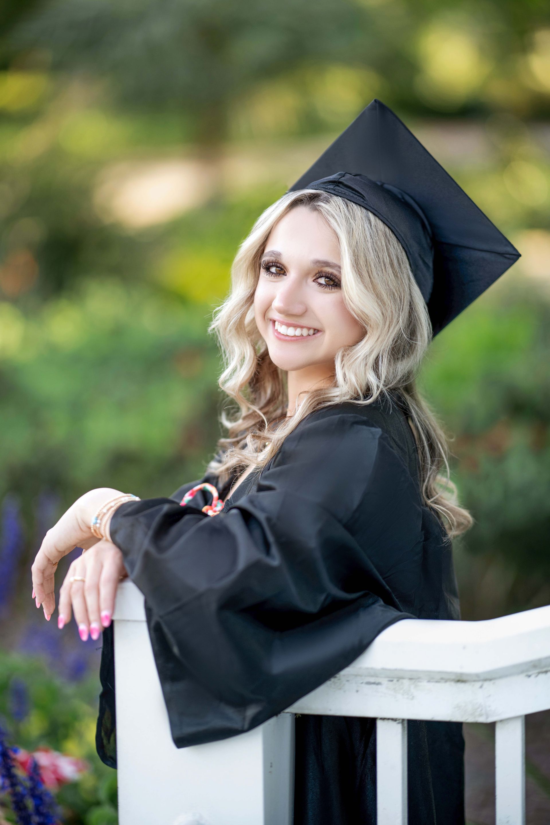 Graduation portrait of a smiling blonde woman in a cap and gown, leaning on a white fence.