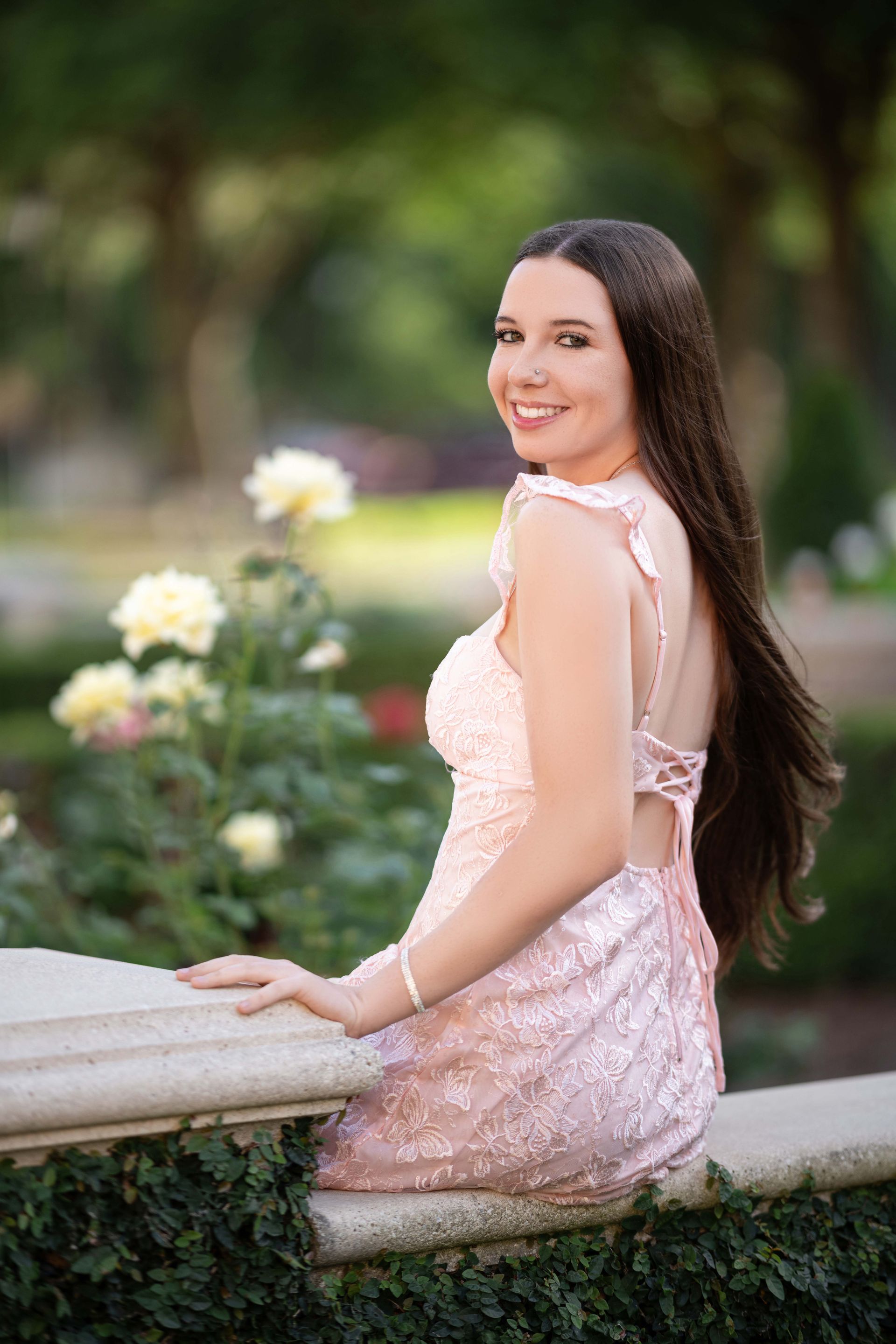 Woman in pink sequined dress, smiles over shoulder at camera while sitting on a stone ledge.