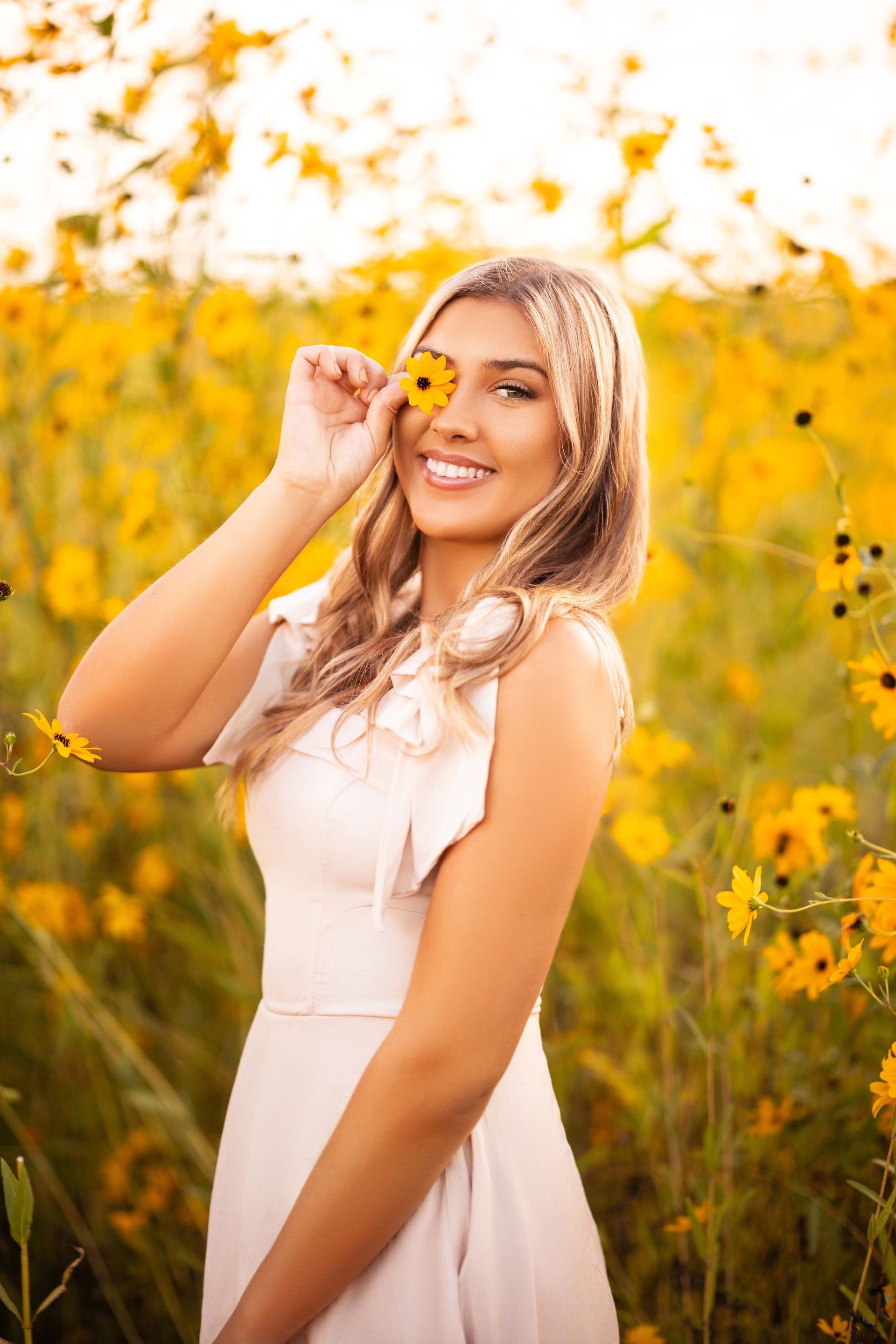 Woman in cream dress holds yellow flower over eye, smiling in field of yellow flowers.