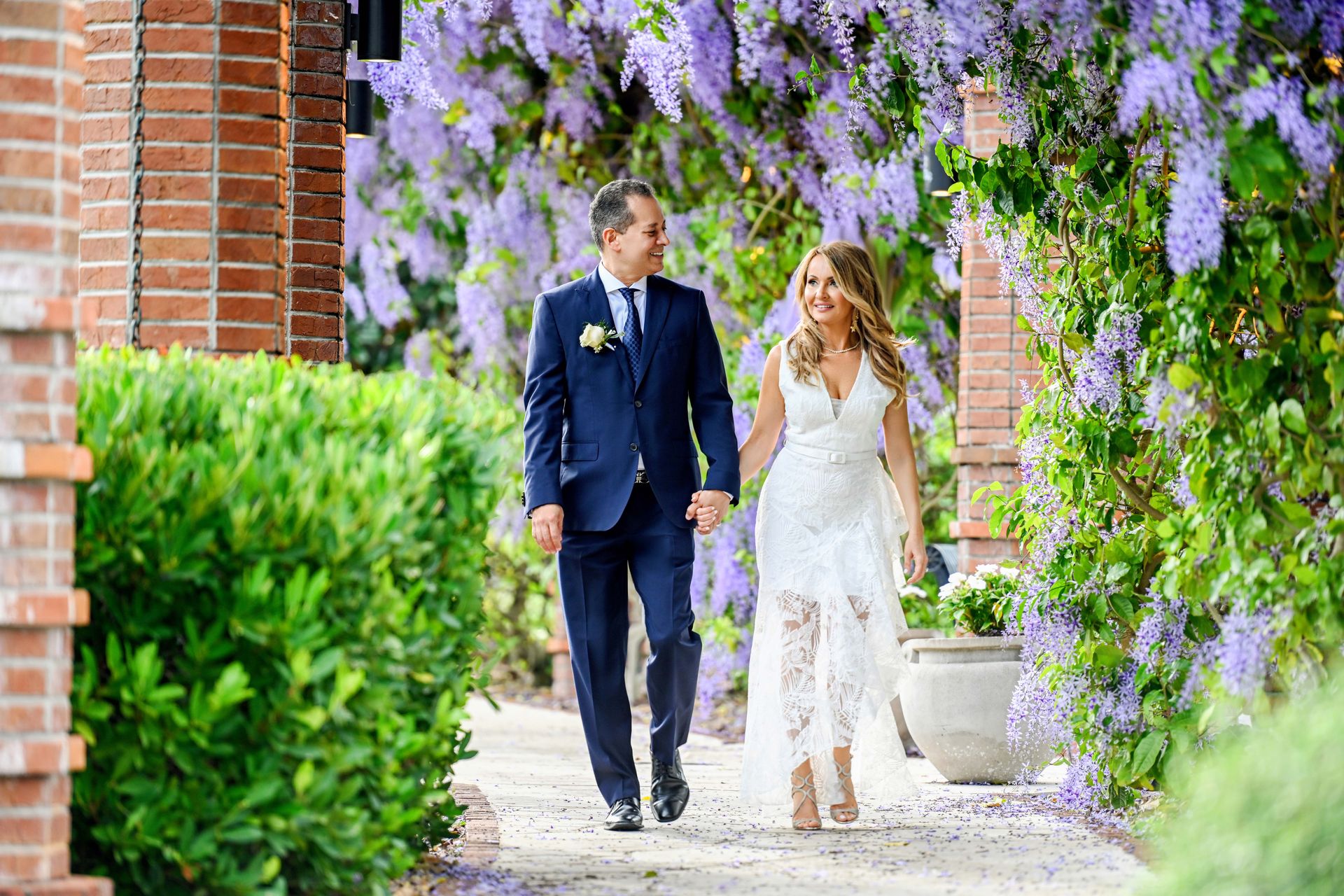 Newly married couple walks hand-in-hand through a garden path lined with wisteria blooms; she in lace, he in a suit.