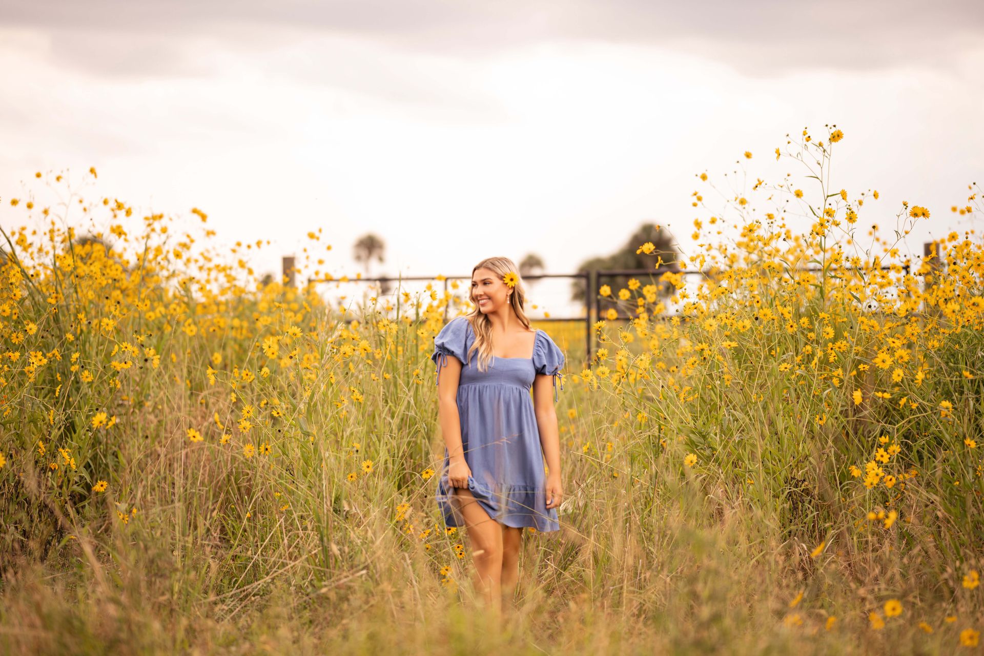 Woman in blue dress smiles, standing in field of yellow flowers.
