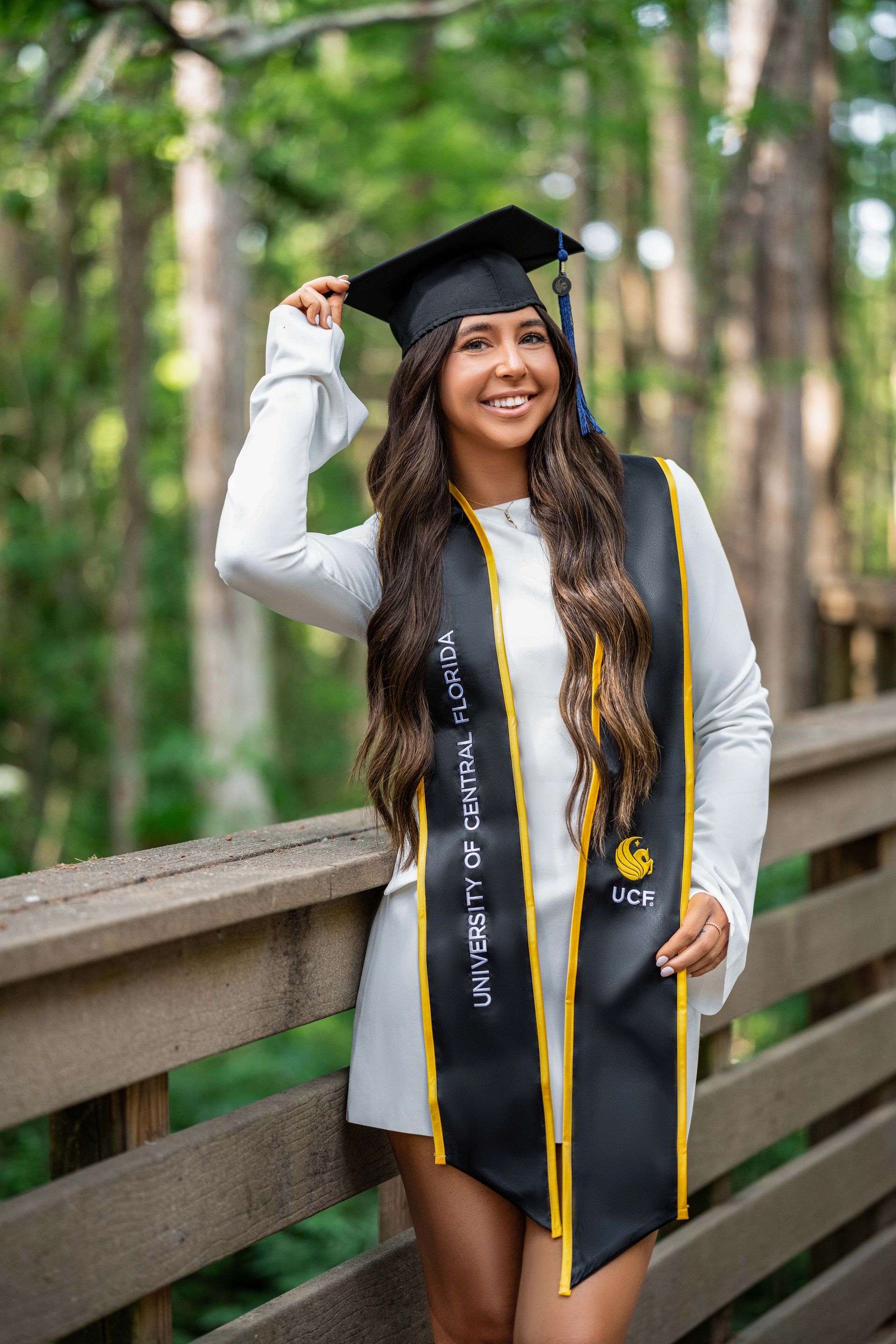Young woman in graduation cap and gown, smiling on a wooden bridge in a forest setting.