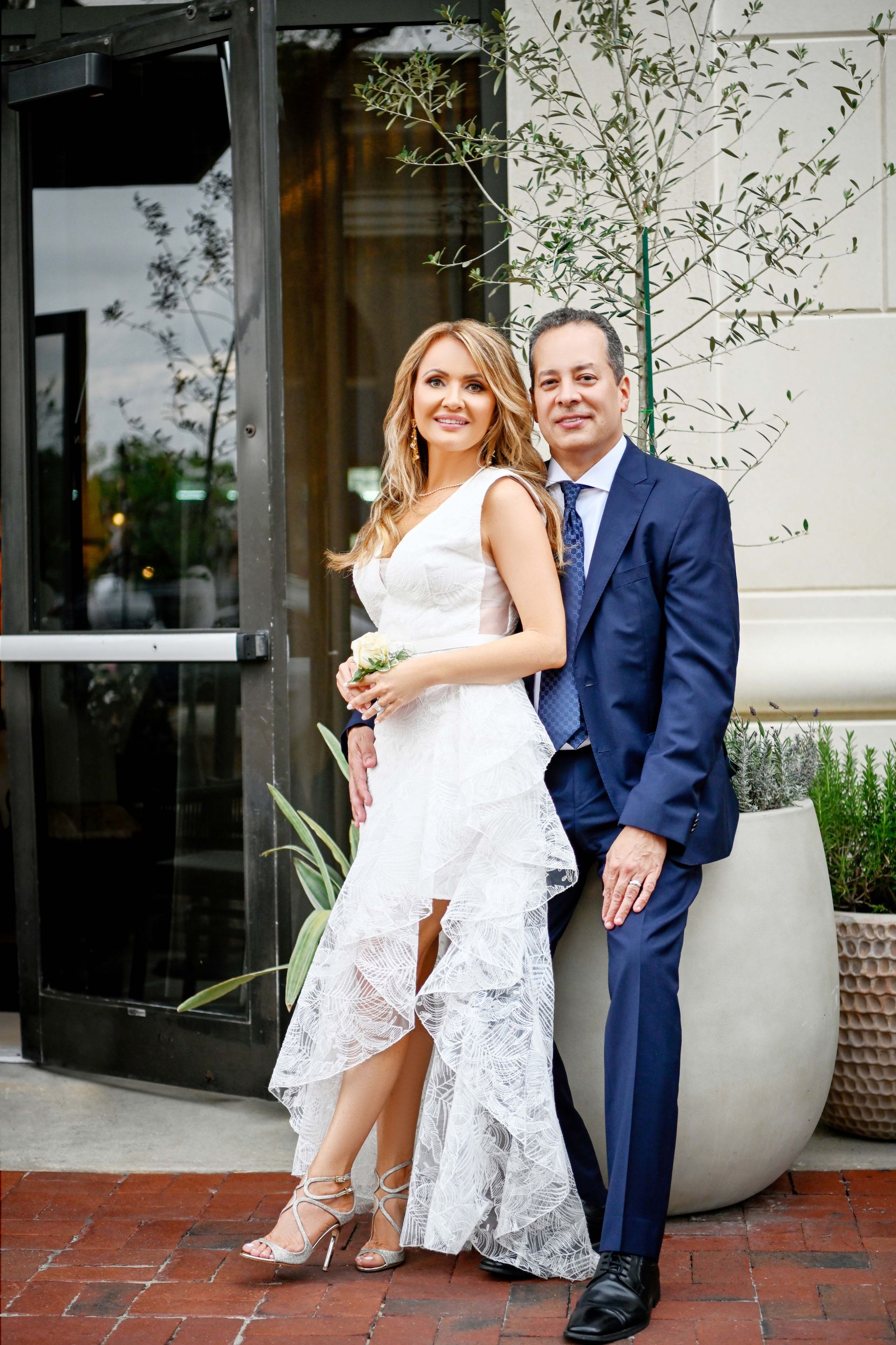 Couple posing outdoors; woman in white dress, man in blue suit, both smiling.