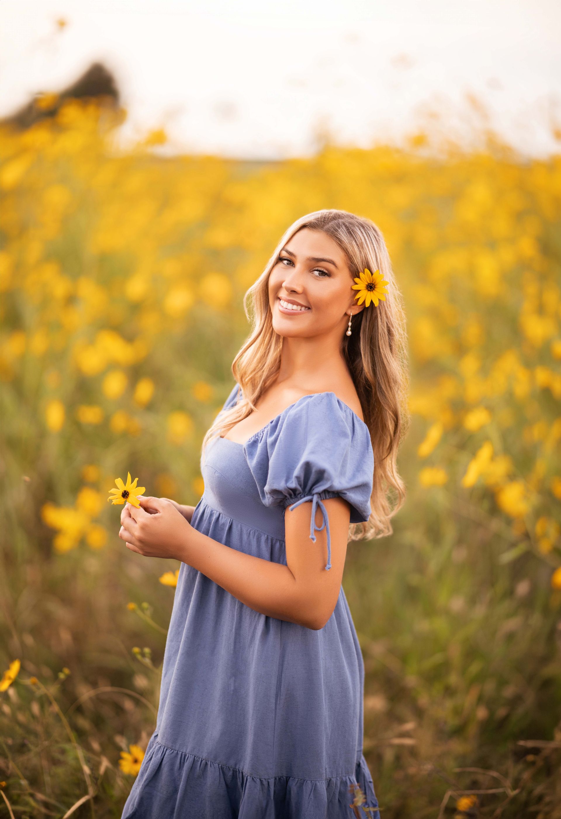 Young woman in blue dress smiling in a field of yellow flowers, holding one.