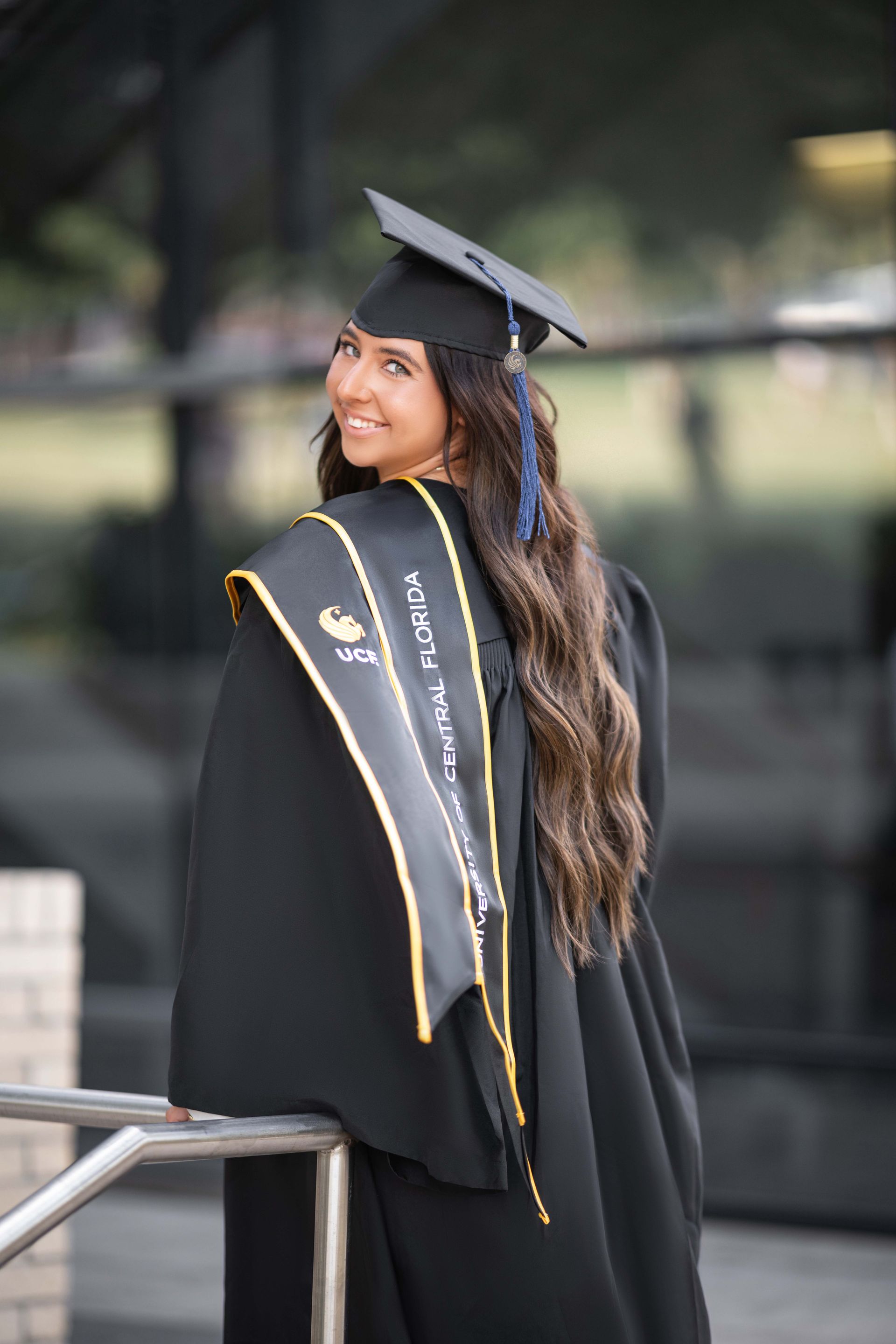 Woman in graduation gown, smiling over her shoulder. Outdoors, near a building. Black gown, gold trim, long dark hair.