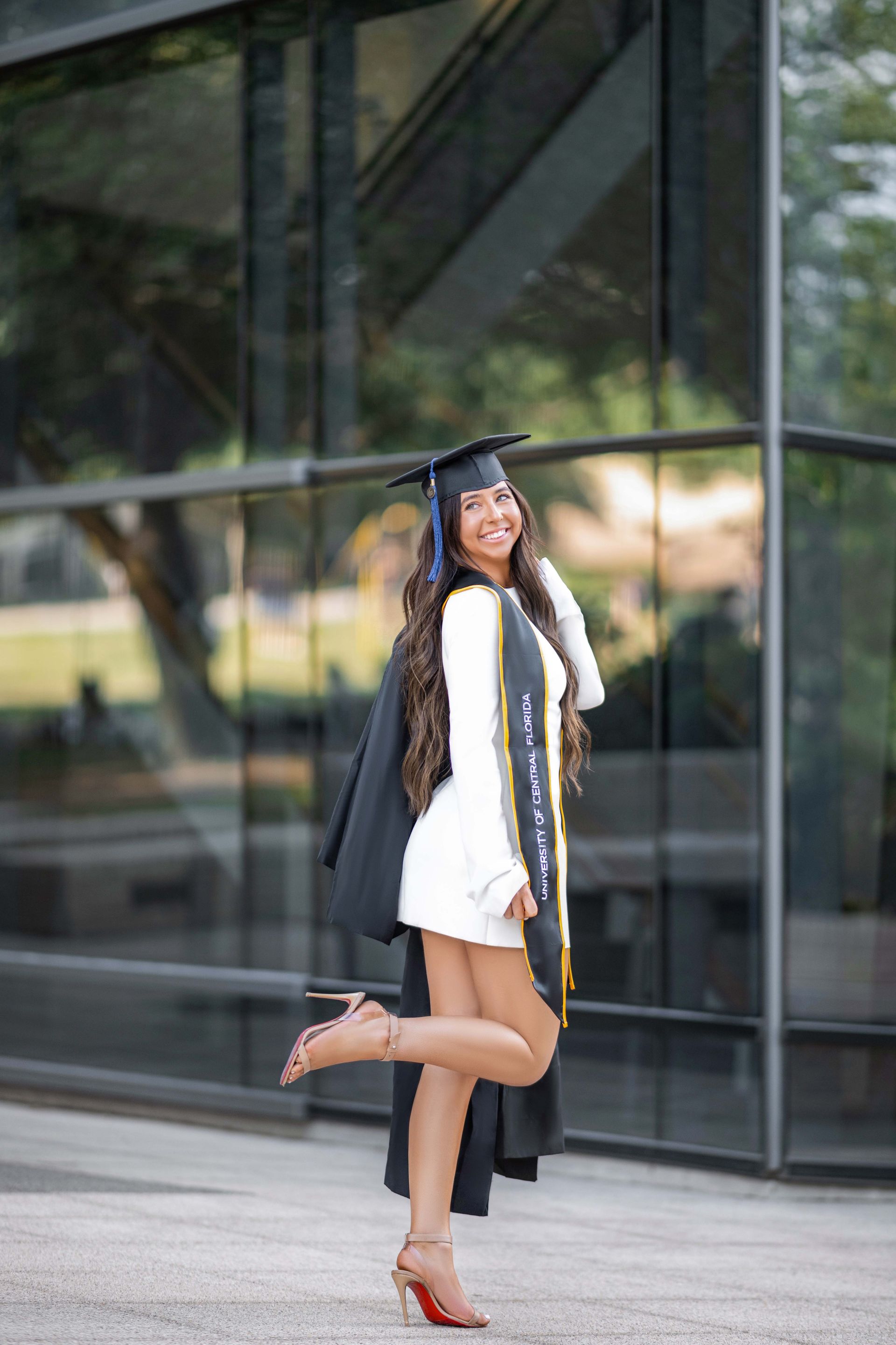 Young woman in graduation attire poses by a building, smiling, leg raised.