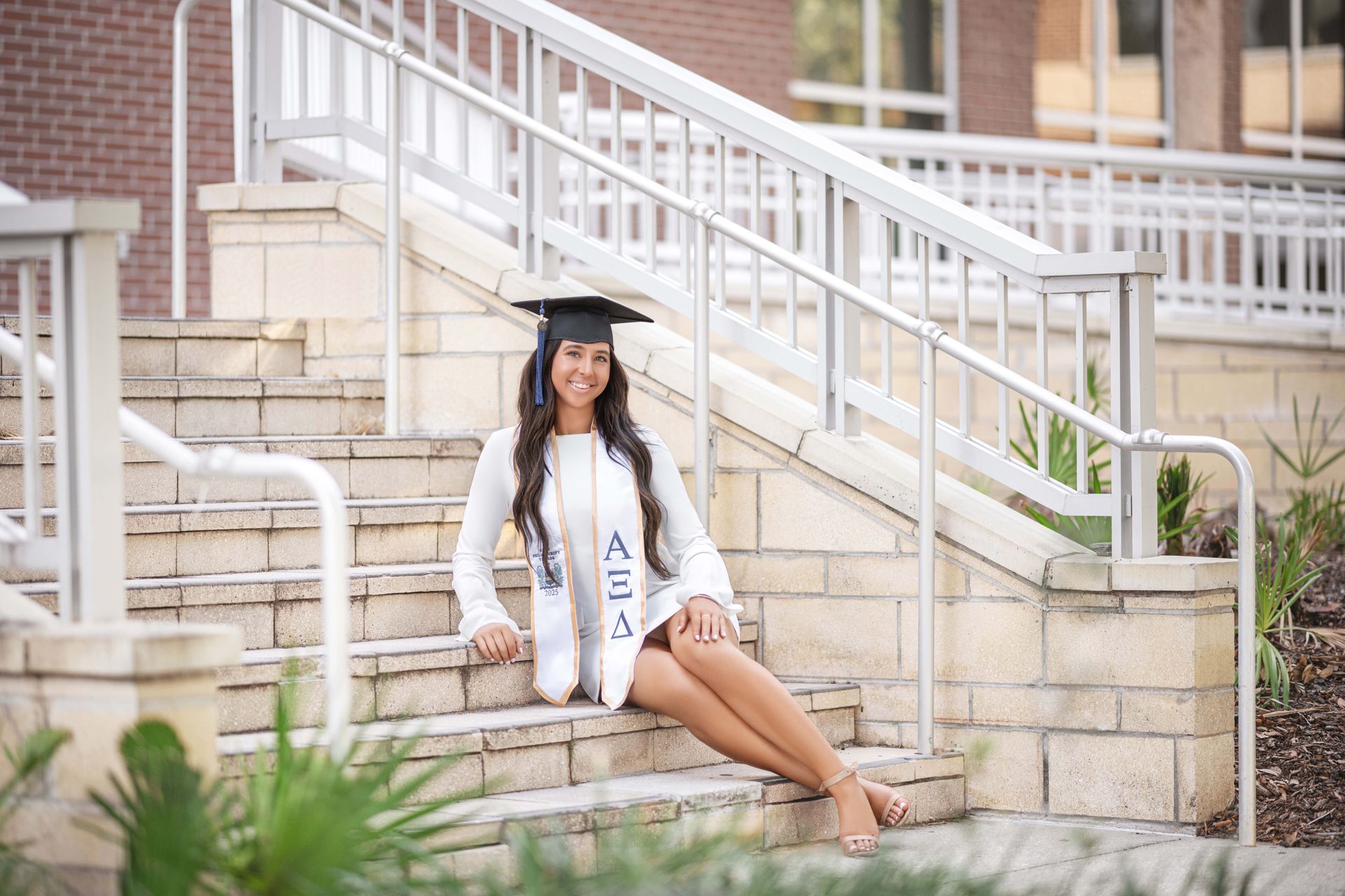 Woman in graduation cap and gown sitting on outdoor steps, smiling.