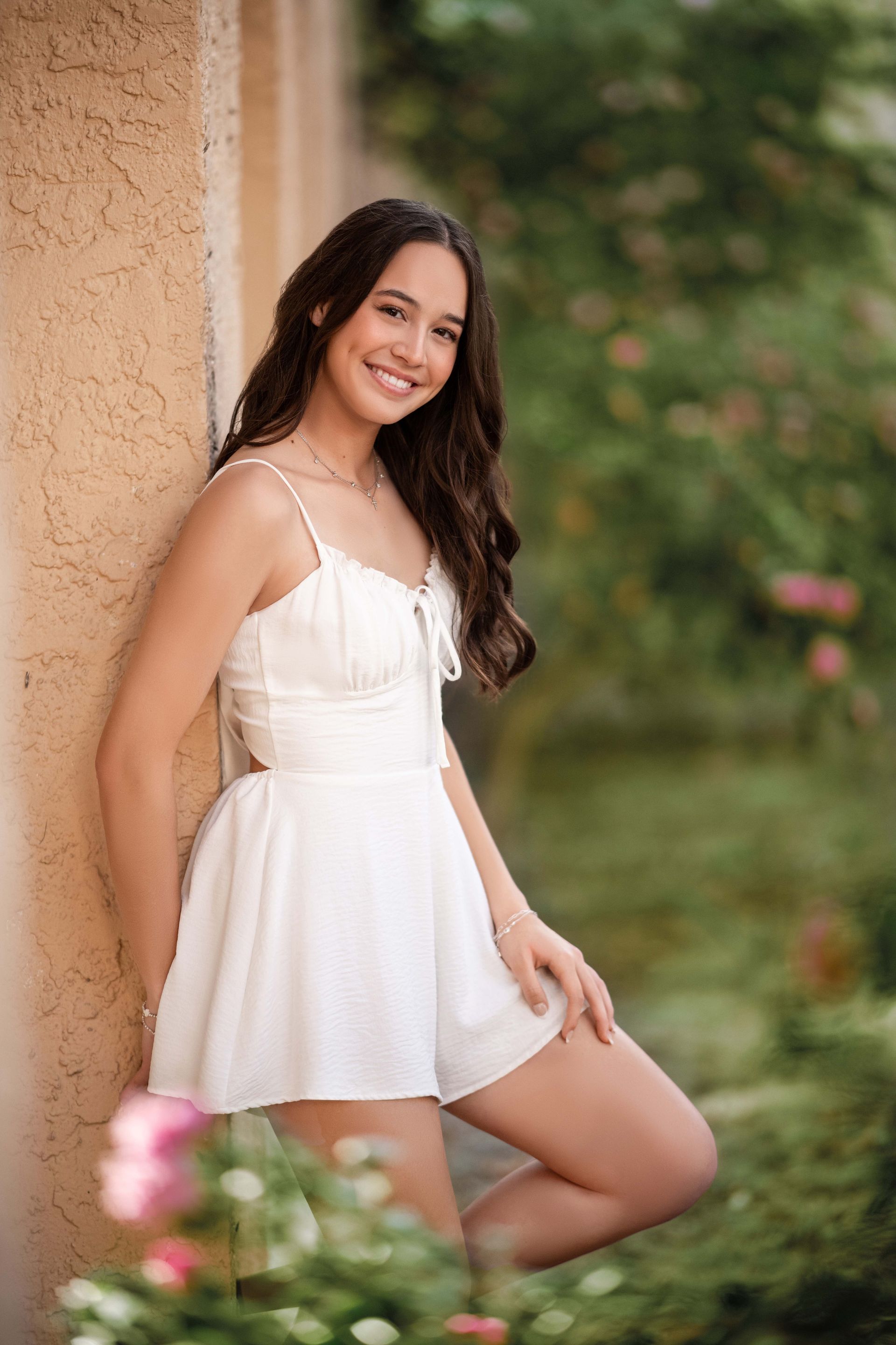 Young woman smiles, leaning against a tan wall, wearing a white romper. Outdoors, near greenery.