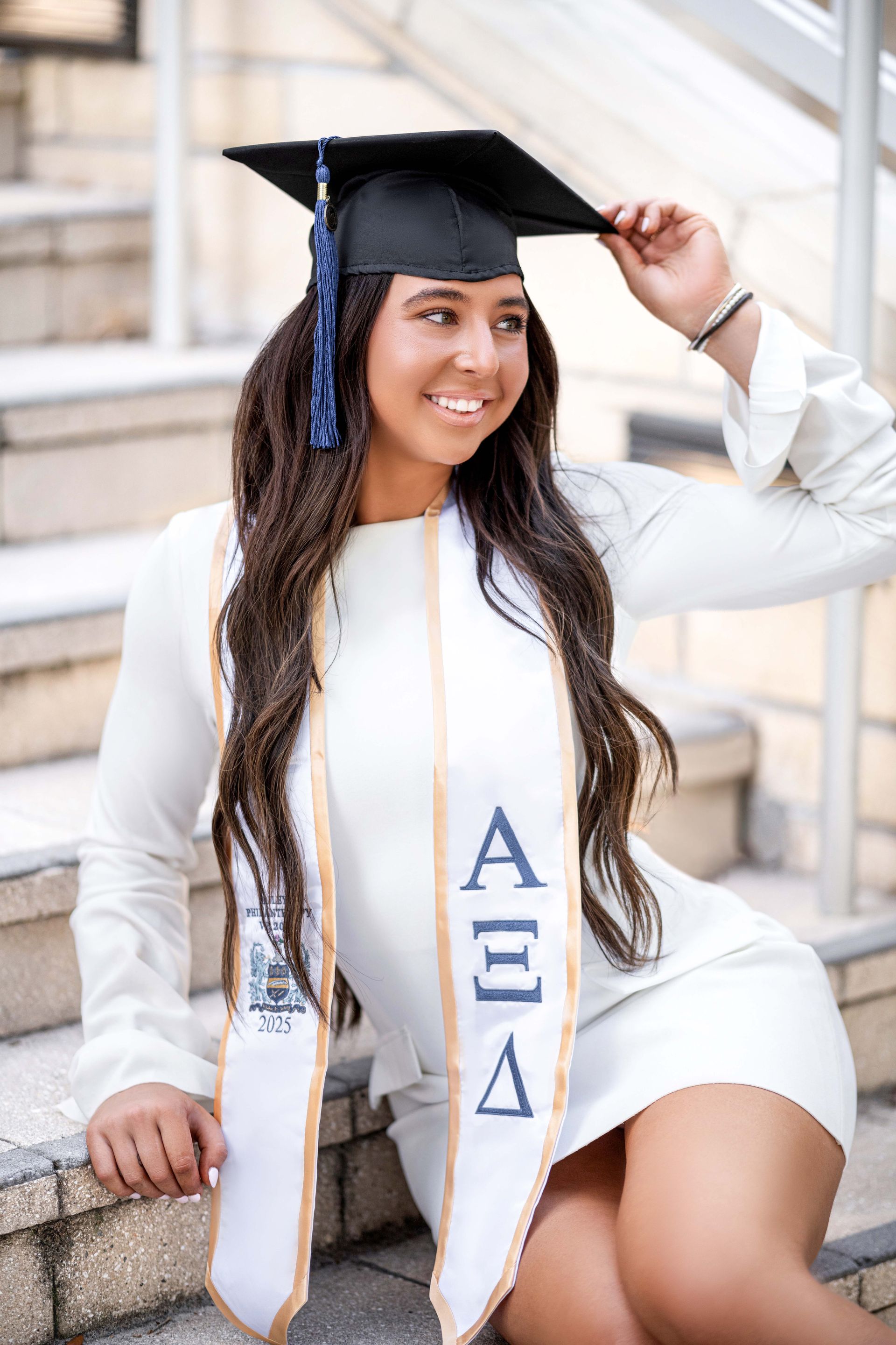 Woman in graduation cap and gown smiling on outdoor steps, wearing a sorority sash.