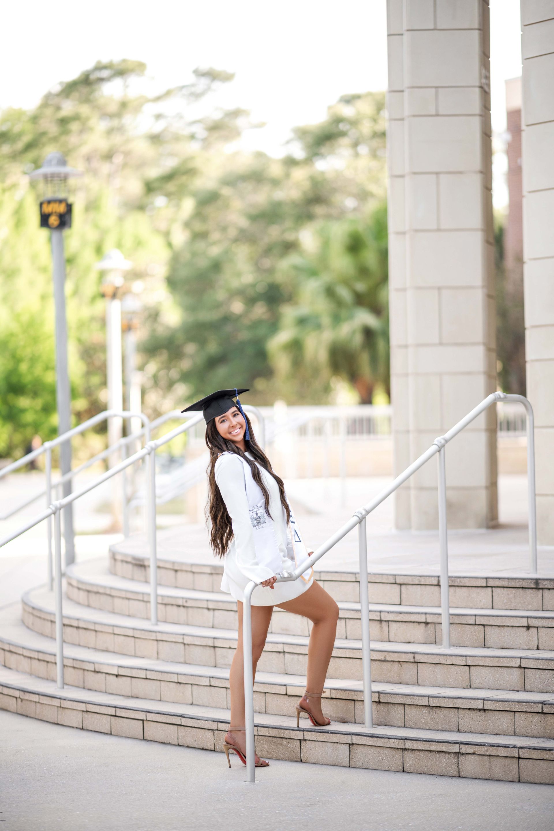 Woman in graduation cap smiles on outdoor stairs. Wearing white, brown heels.