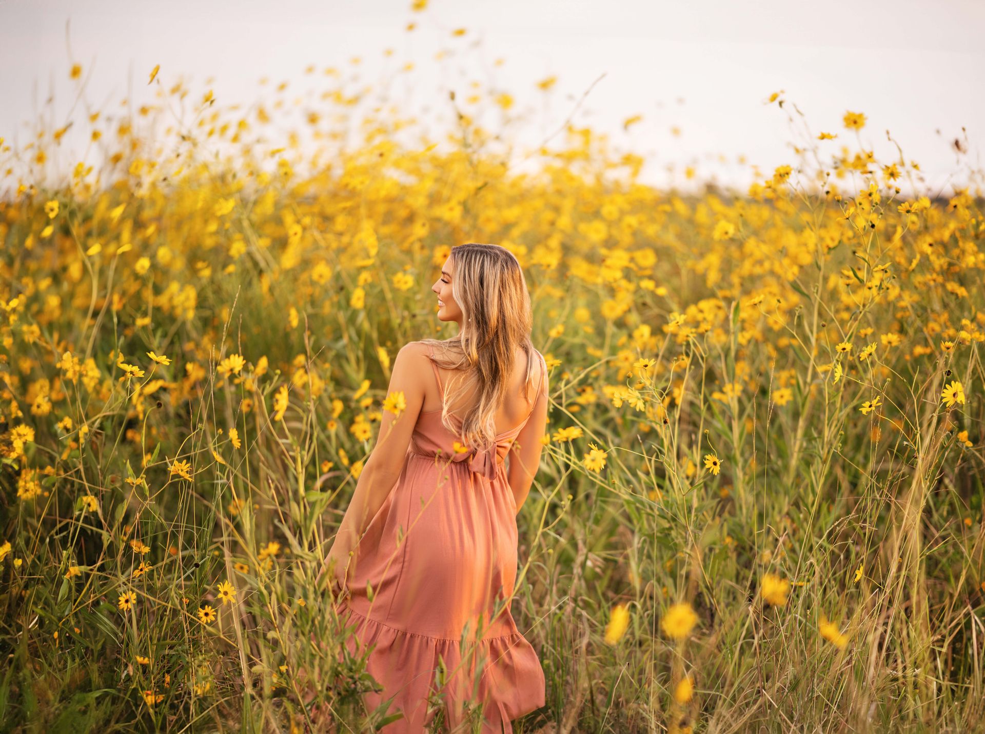 Woman in a flowing peach dress, turns to face the camera in a field of yellow wildflowers, sunlight.