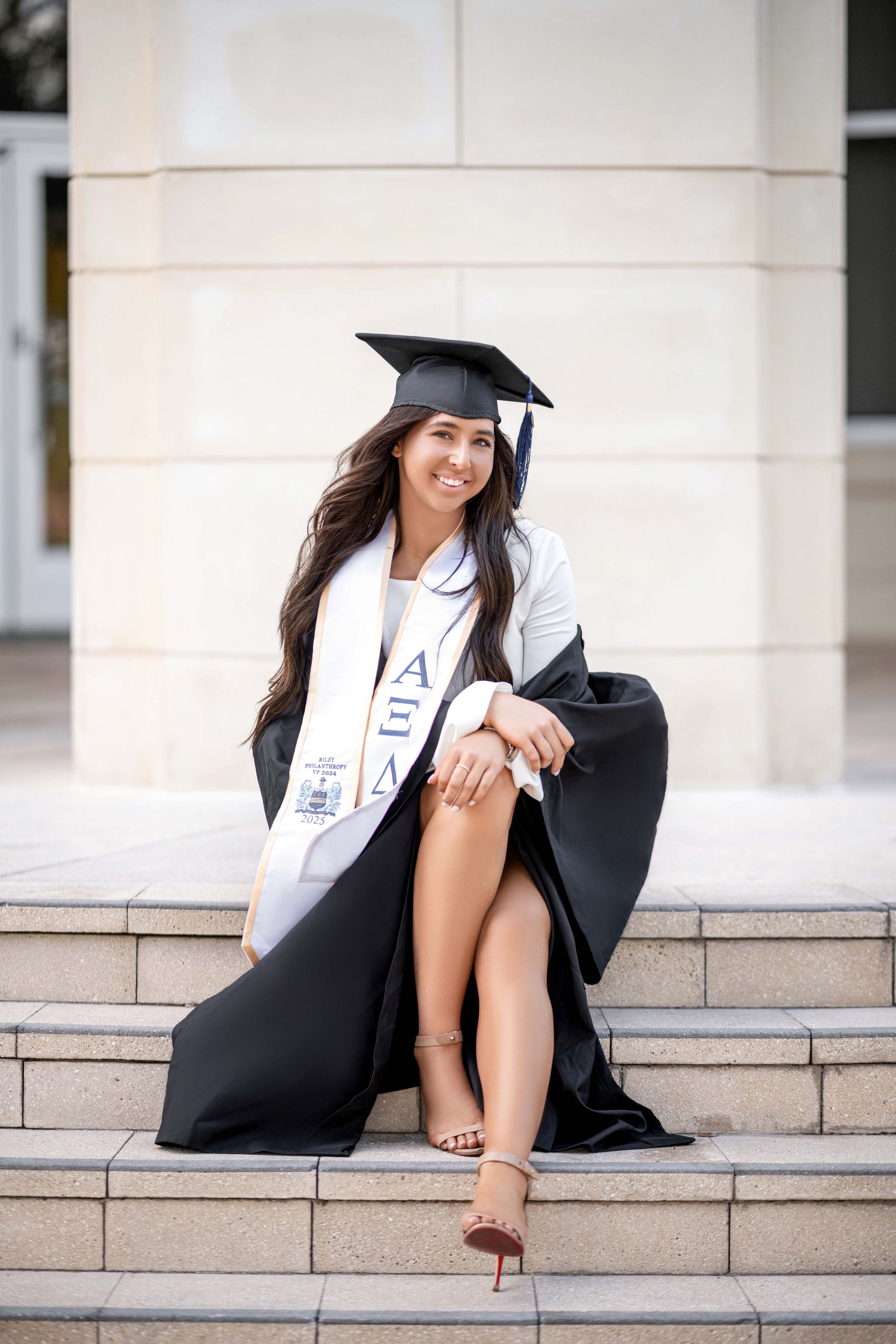 Woman in graduation cap and gown, smiling and seated on steps.