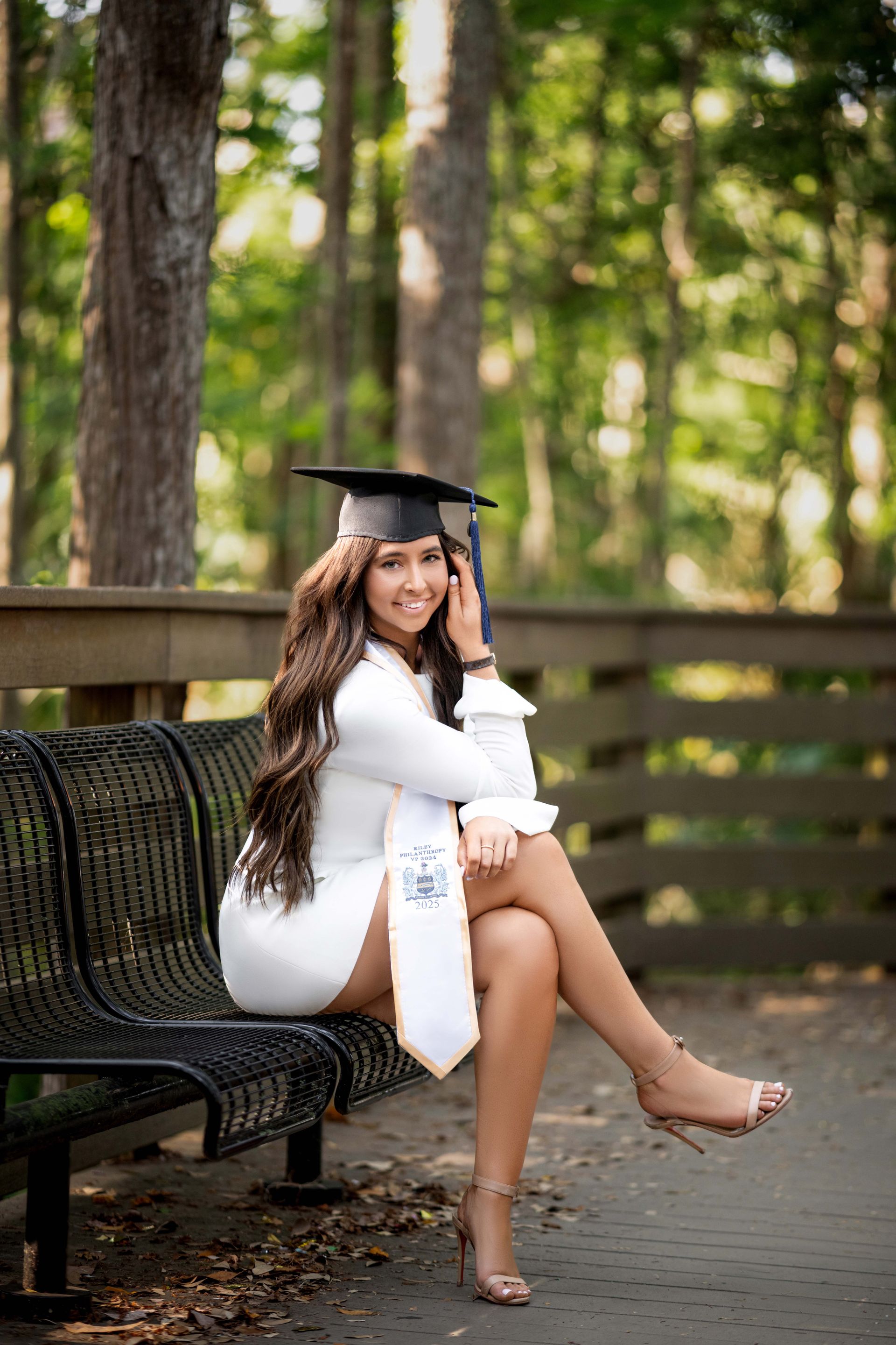 Woman in graduation attire smiles on a bench in a wooded area. She wears a white dress, cap, and sash.
