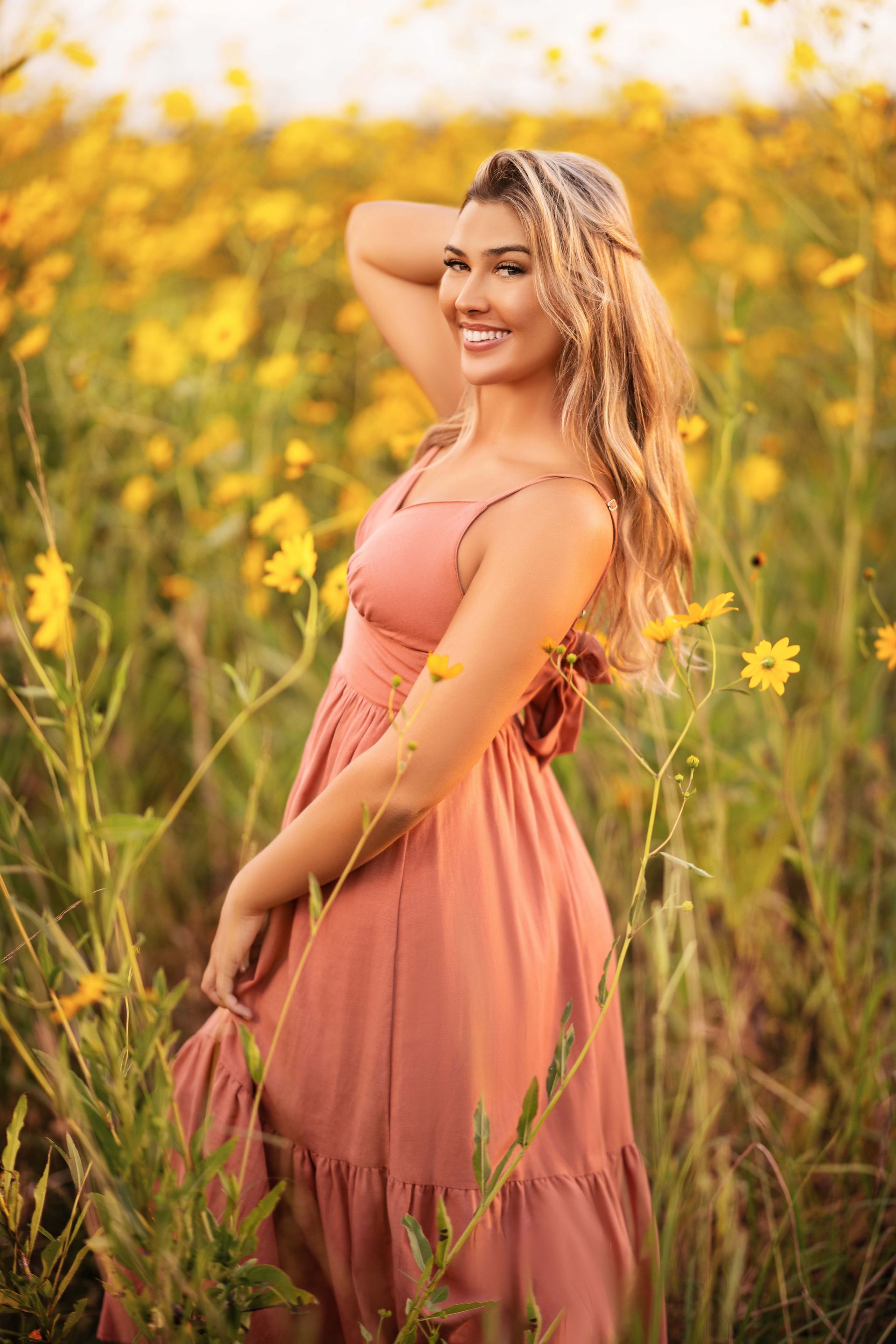 Woman in a pink dress smiles, standing in a field of yellow flowers, arm raised, golden hour lighting.